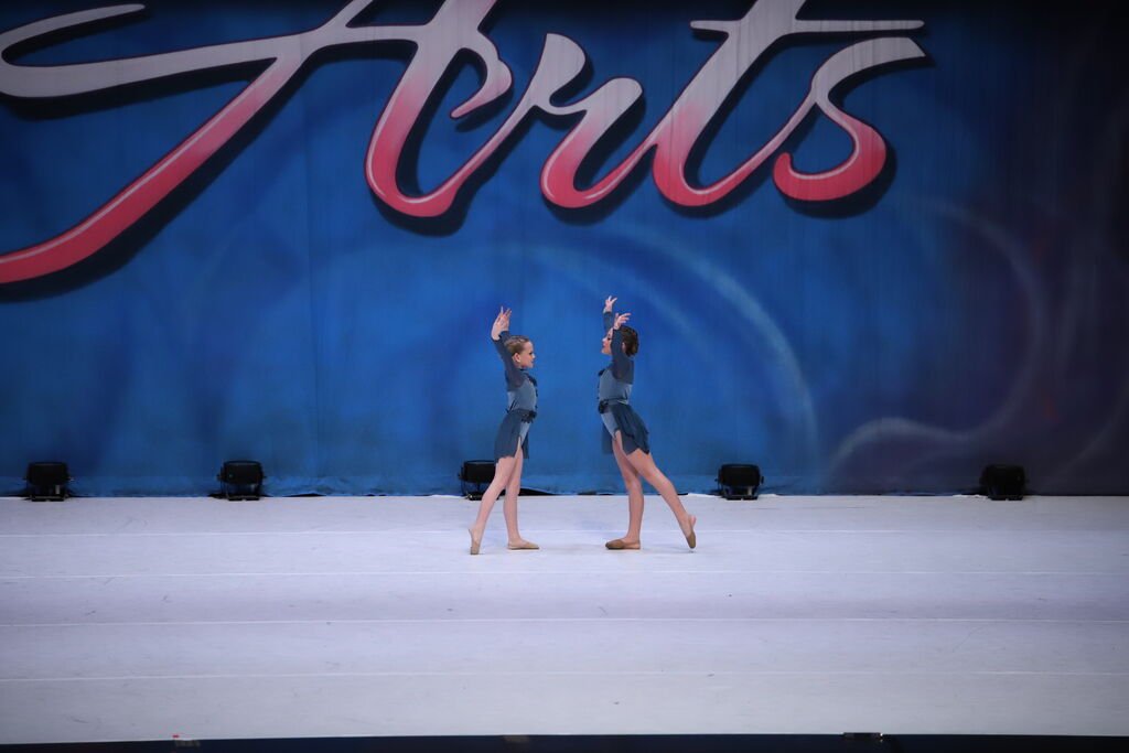 Two young girls perform a dance routine on stage in front of a large blue backdrop with the word 'Truist' in stylized text. They are dressed in coordinated dance costumes and are engaged in a synchronized dance move.