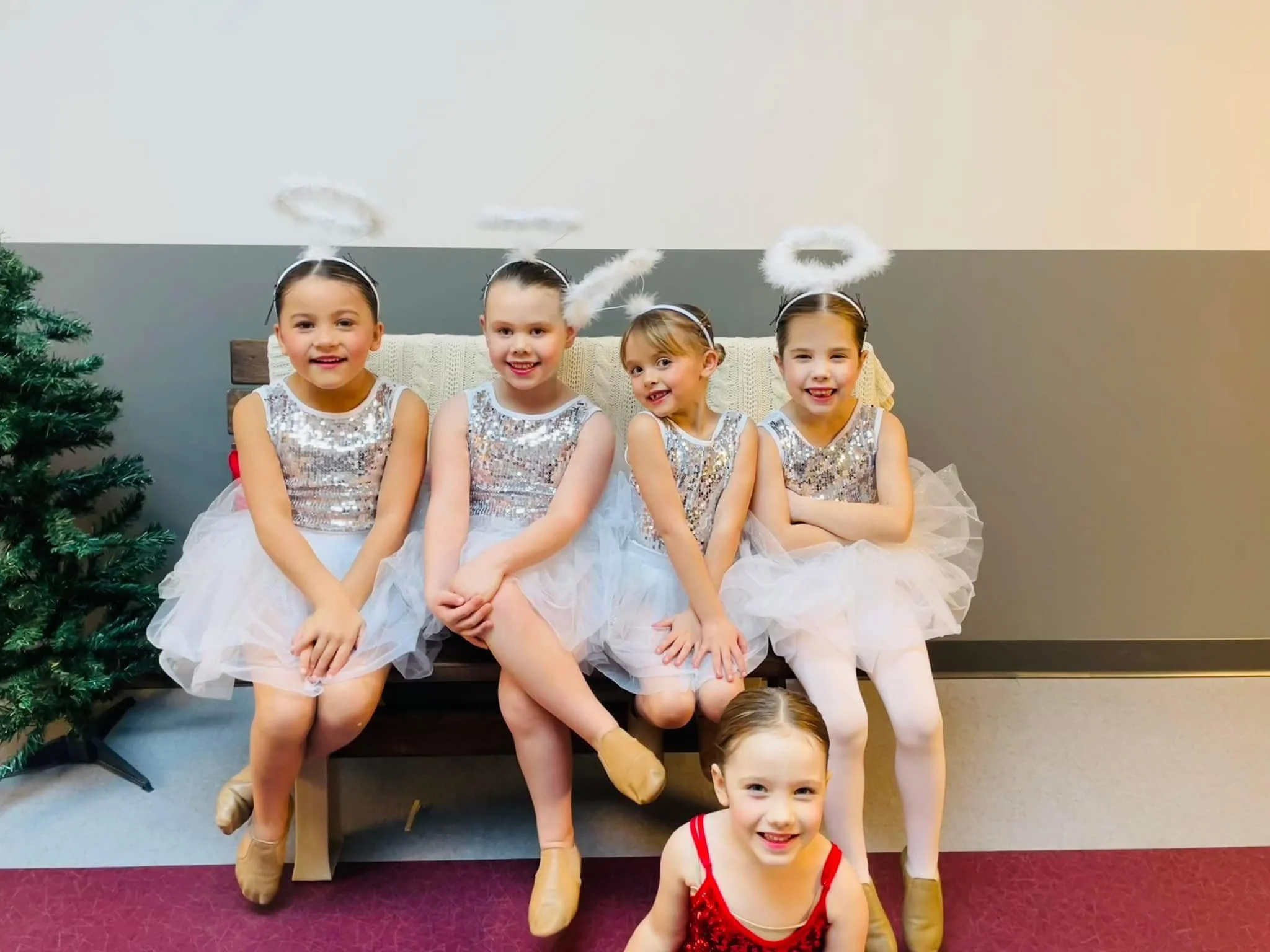 Five young girls dressed as angels with white dresses, sequined bodices, halos, and ballet slippers, sitting on a bench with one girl in a red dress kneeling on the floor in front of them. A Christmas tree is partially visible to the left.