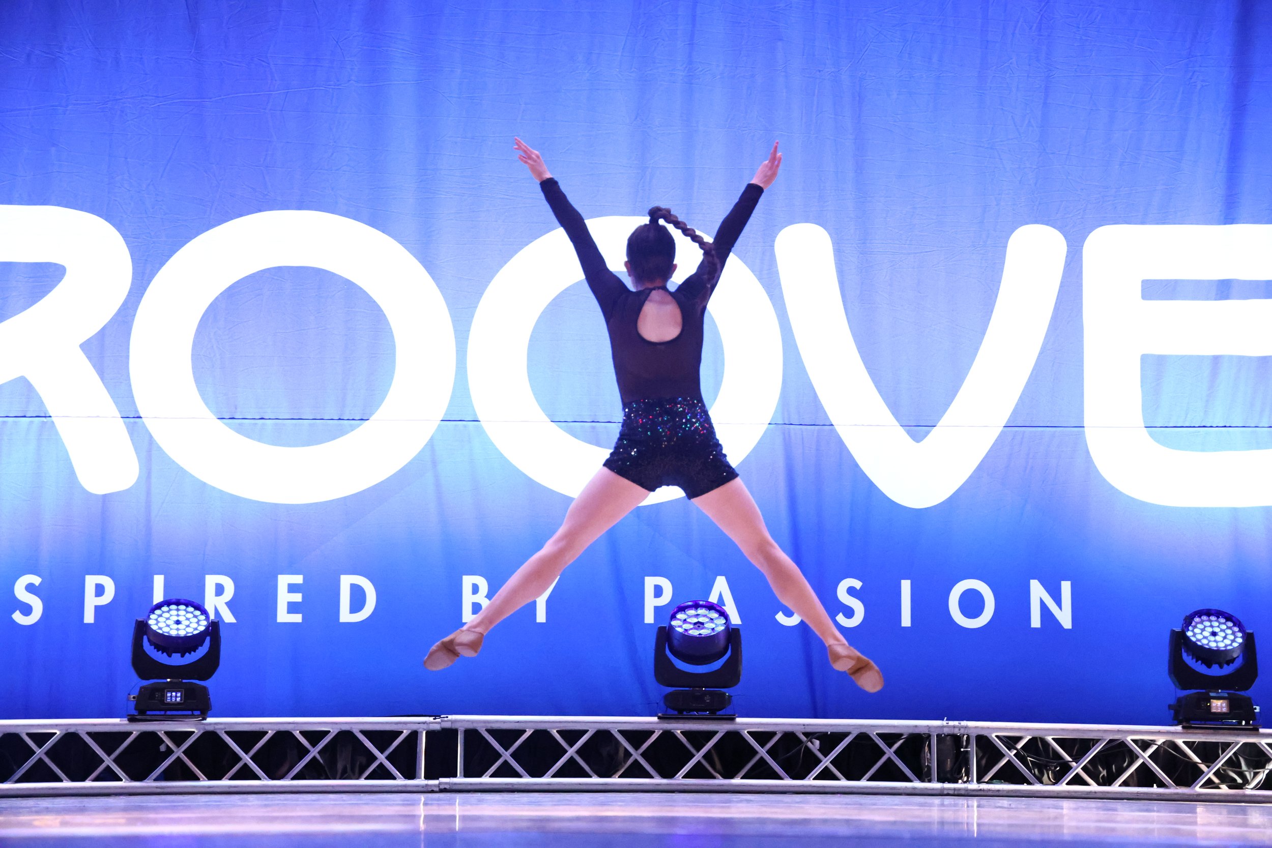 A young female dancer performing a leap on stage with a blue backdrop that says 'ROOV' and 'SPIRITED BY PASSION'. She is wearing a black top with an open back and sequined shorts.