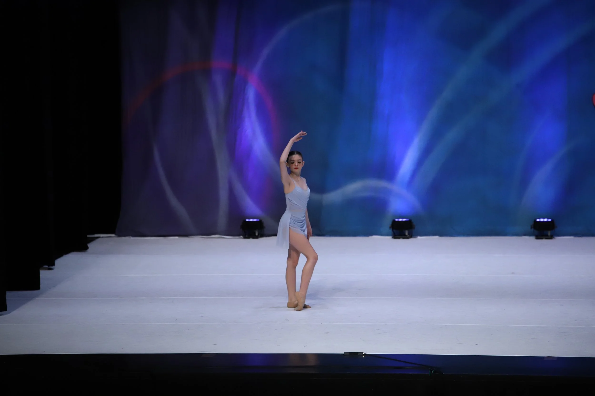 A young woman performing a ballet dance on stage with a blue and purple light backdrop.