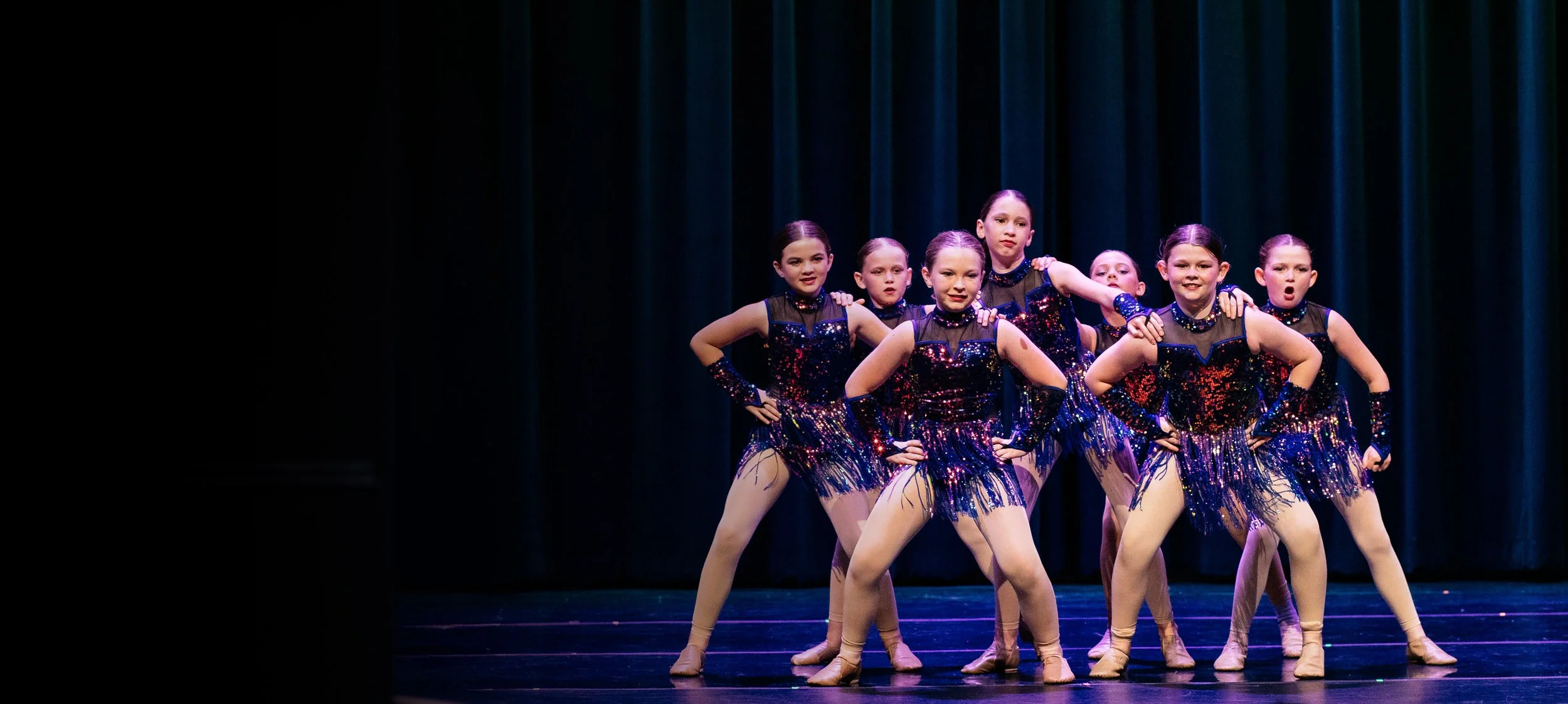 A group of young ballerinas performing on stage in shiny costumes with black mesh tops and colorful skirts, posing with hands on hips against dark blue curtains.