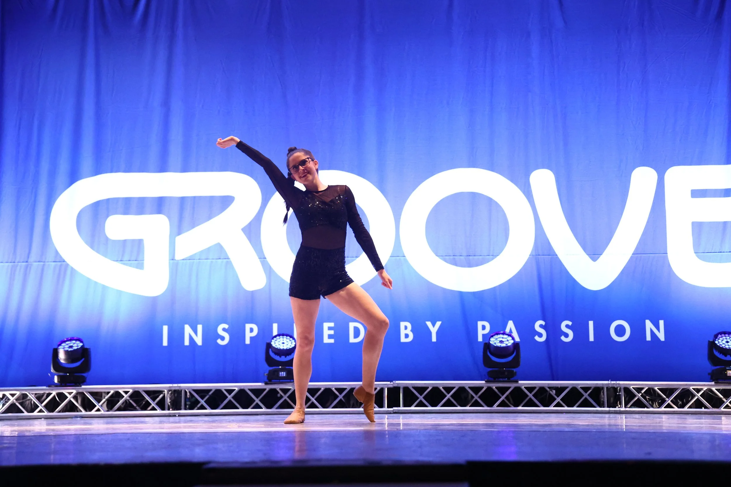 A young woman performs a dance pose on stage at Groove event with large backdrop reading "GROOVE" and "INSPIRED BY PASSION".