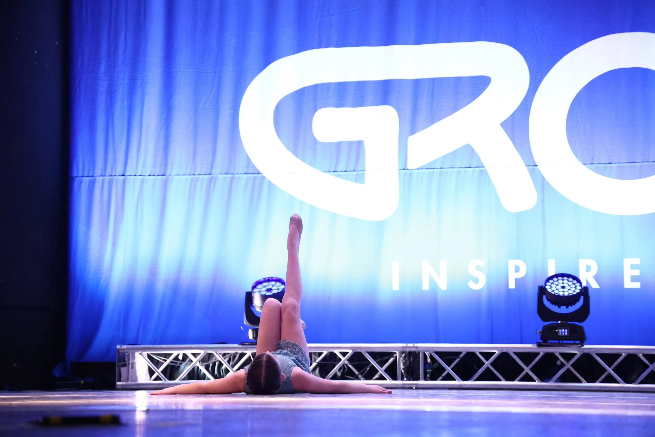 A young girl performing a dance pose in front of a large blue curtain with the words 'GRO INSPIRE' displayed on it, on a stage lit with blue and purple lights.