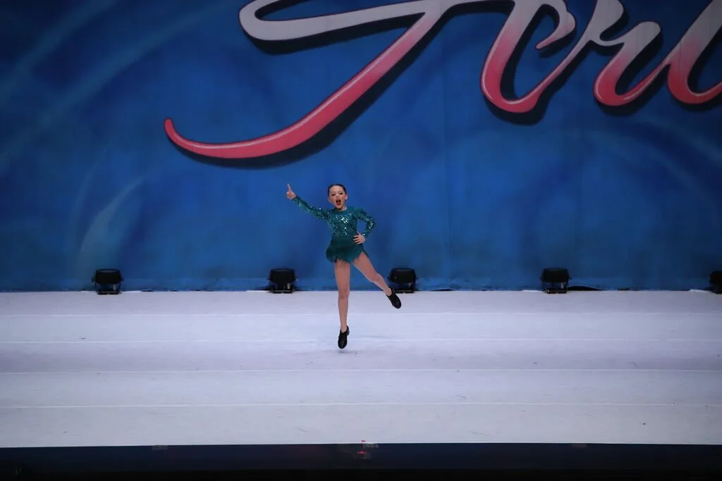 A young woman performing an ice skating routine on a stage with a blue backdrop and the word "Toy" written in large cursive letters.