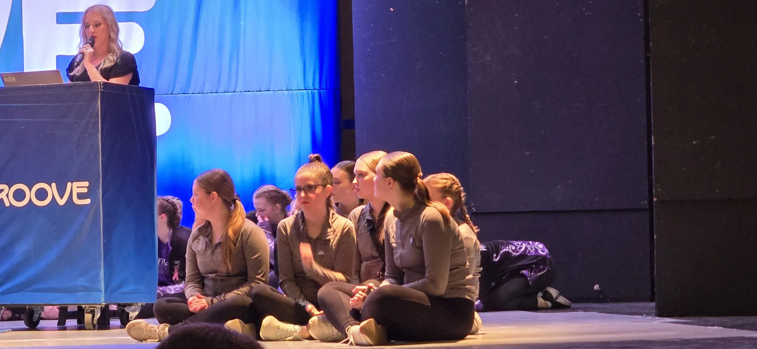 A group of young women sitting on the floor in front of a stage, listening to a woman speaking at a podium against a blue backdrop with the word 'GROOVE' on it.