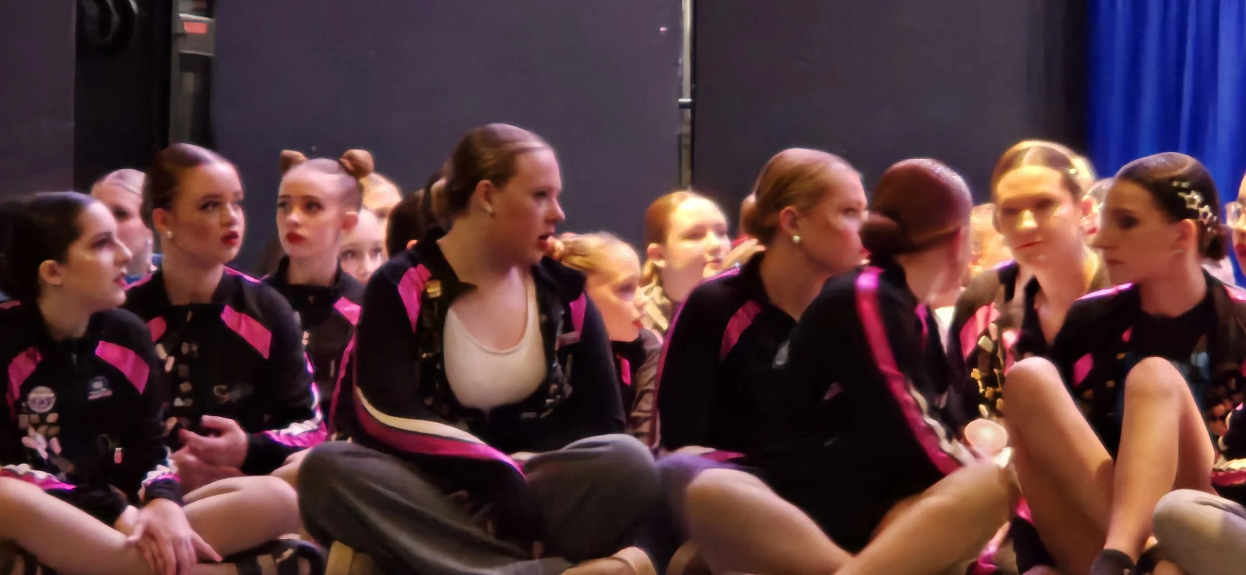 Group of young female gymnasts sitting on the floor, some talking and some listening attentively, in matching black and pink team uniforms.