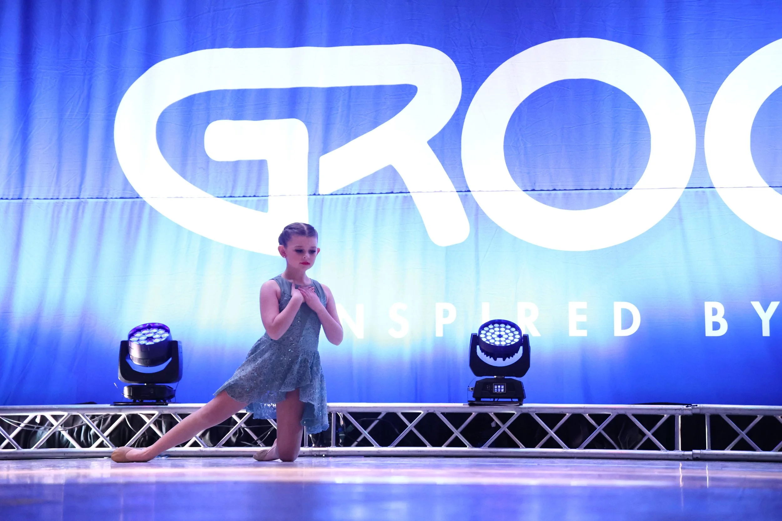 Young girl in a dance costume performing a dance move on stage with a large blue backdrop and stage lights.