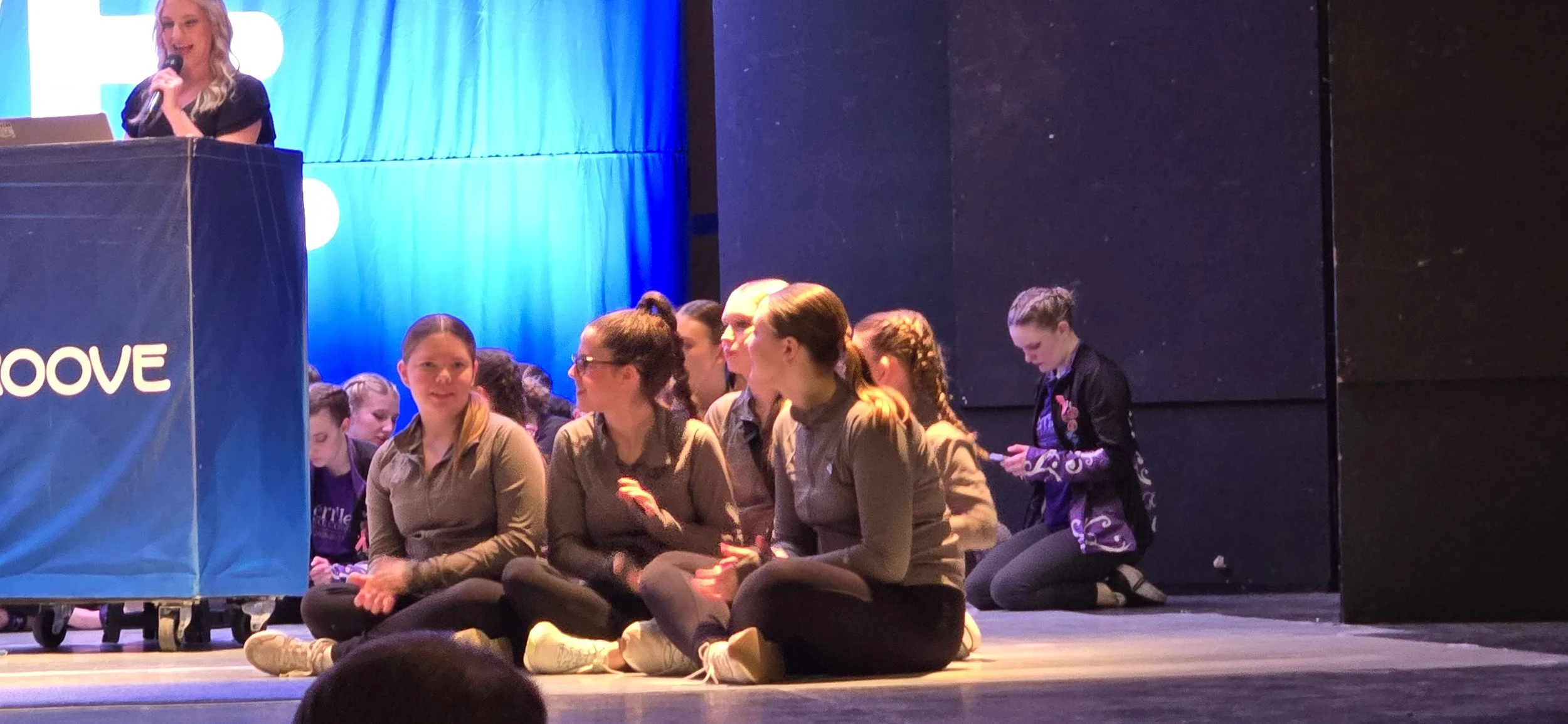 Group of young women sitting cross-legged on stage, with one woman standing at a podium speaking into a microphone, in front of a blue curtain backdrop.