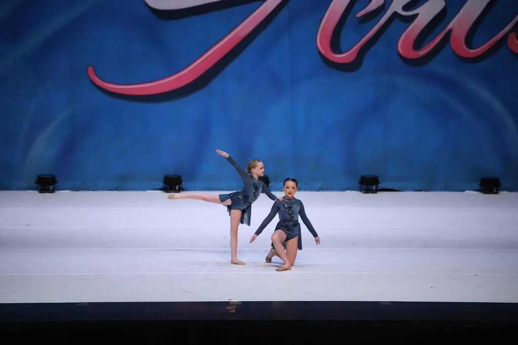 Two young girls in blue dance costumes perform a routine on stage, one standing with an arm out and leg extended, the other kneeling, during a dance competition.