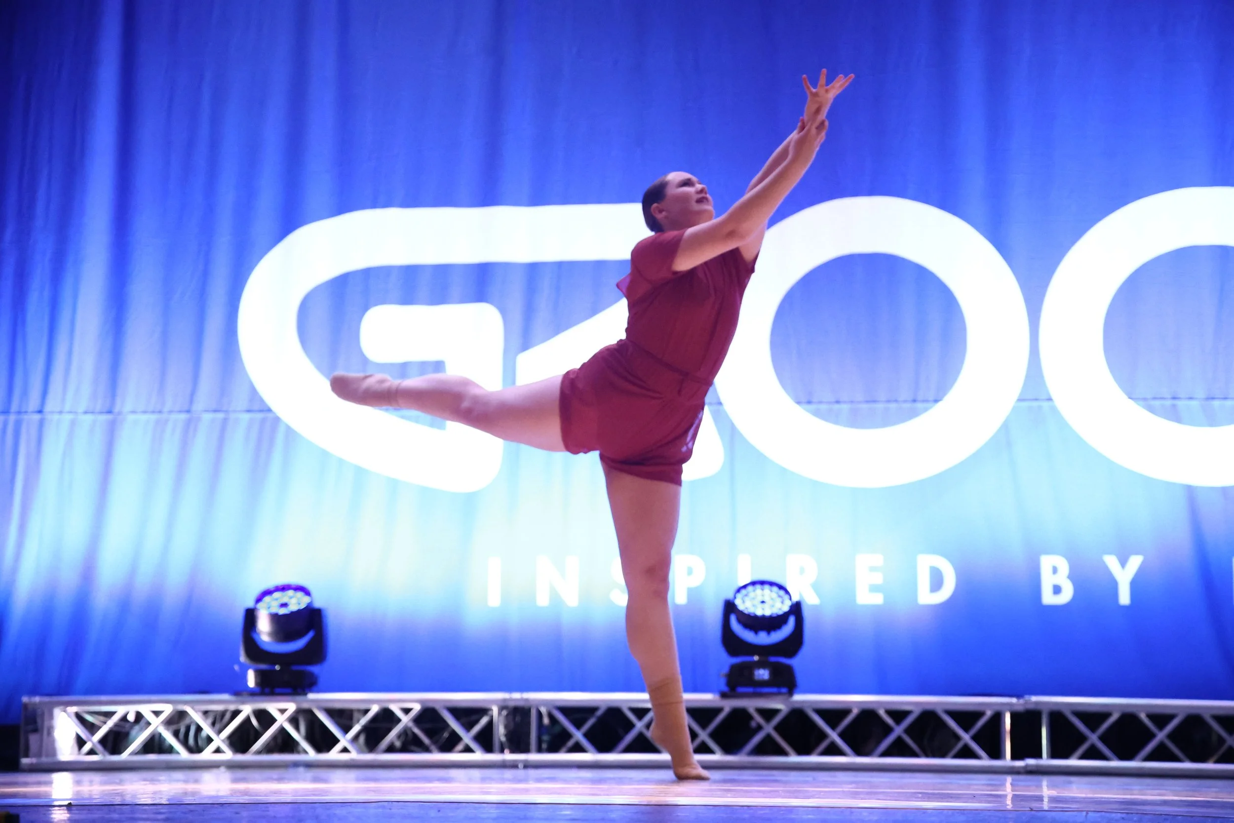 A woman performing a dance or gymnastics move on stage, standing on one leg with the other extended behind her, and arms reaching upward. She is in front of a blue backdrop with large white text and stage lights.