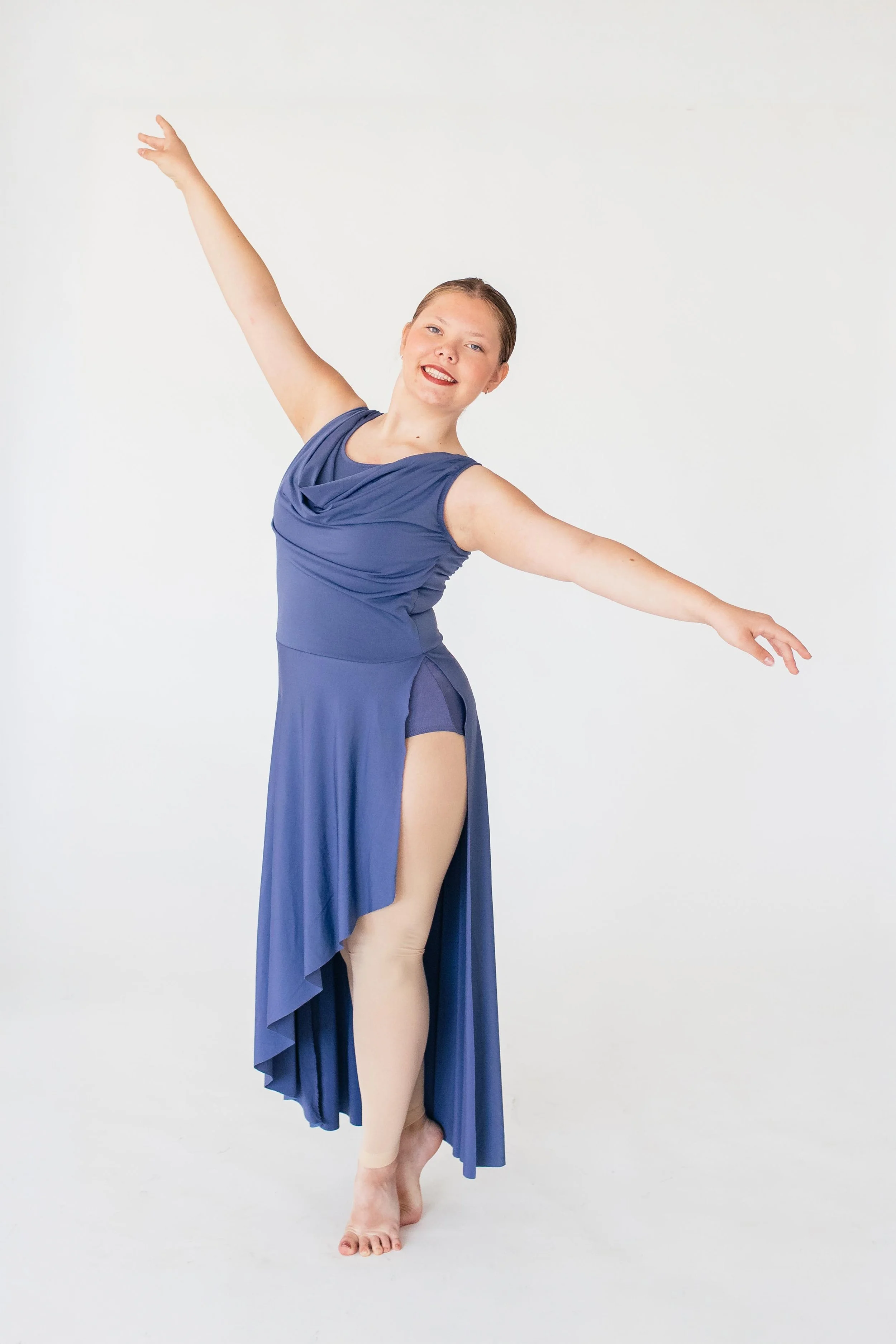 Ballet dancer in a blue dress, holding a pose with arms extended, standing on one leg with the other leg slightly bent, against a white background.