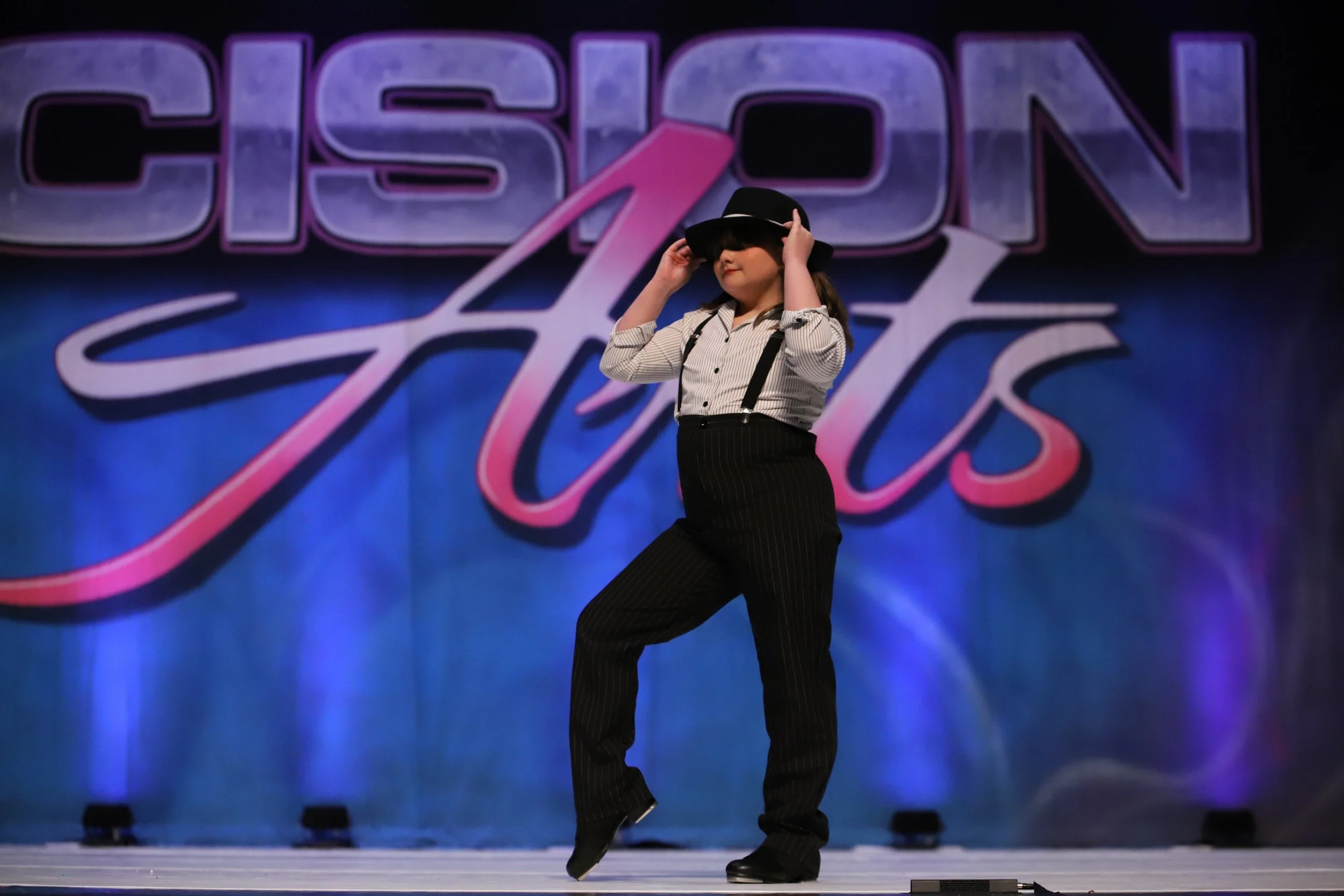Young girl performing a dance routine on stage at a dance competition, wearing a hat, suspenders, and black dress pants, with a large sign reading 'Cison Arts' in the background.