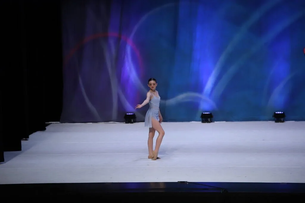 A young female dancer in a light blue costume performing on a stage with side curtains and a colorful, abstract background.