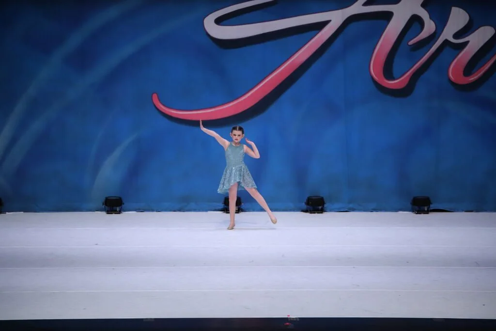Young girl wearing a sparkly blue dance costume performing a dance move on stage, with a blue backdrop and stage lights.