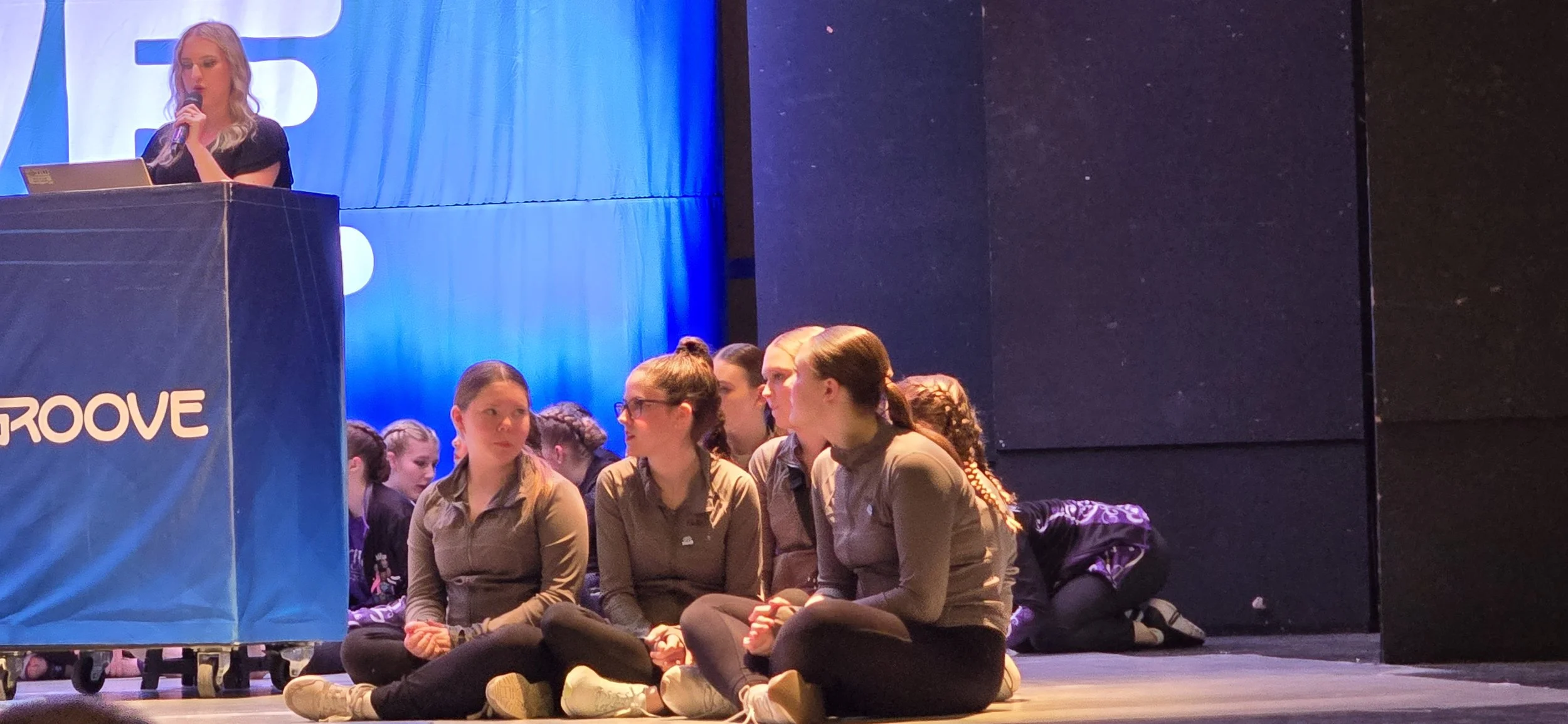 A woman is speaking into a microphone at a podium on stage. In front of her, a group of young women are sitting cross-legged on the floor, some engaged in conversation while others look on, in a dimly lit auditorium with blue lighting and a dark back