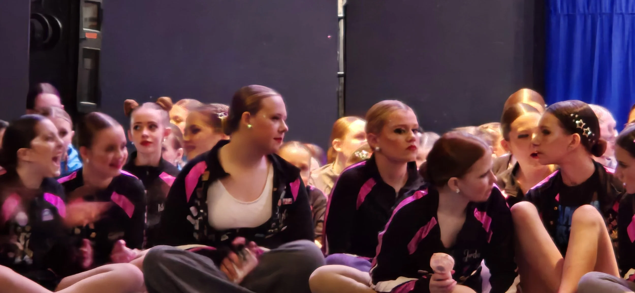 Group of young girls sitting on the floor in a dance studio, dressed in athletic wear, appearing to wait or watch something.