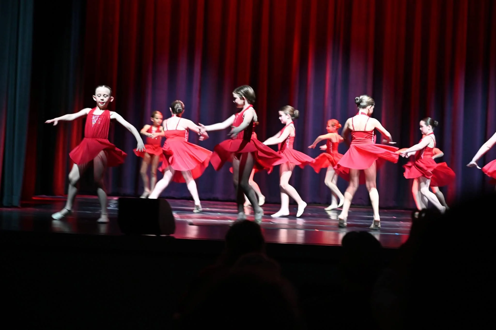 Young girls performing a ballet dance in red costumes on stage with a dark red curtain background.