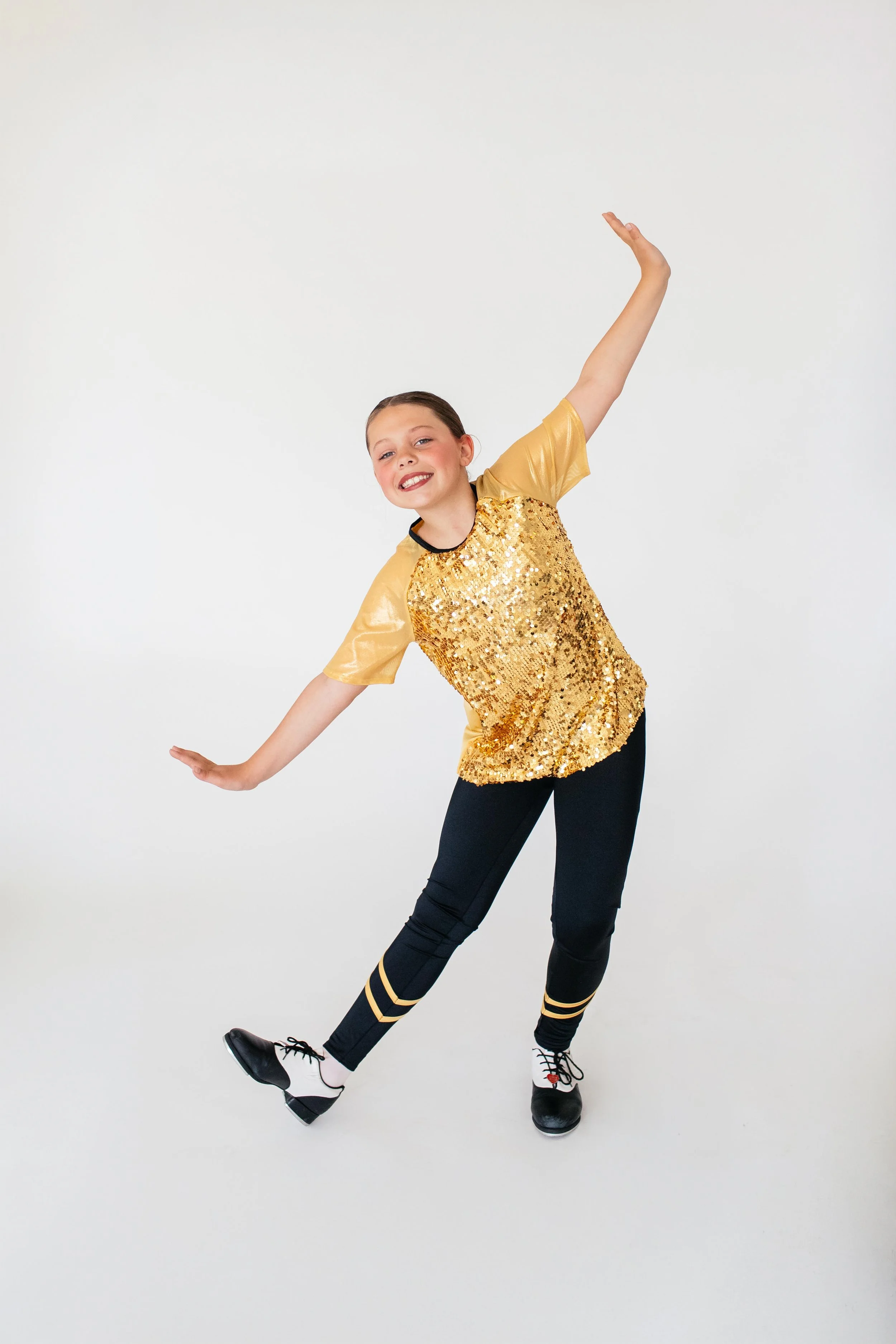 A young girl with dark hair in a bun wearing a gold sequin top, black pants with gold stripes, and white sneakers posing with arms outstretched and smiling against a plain white background.