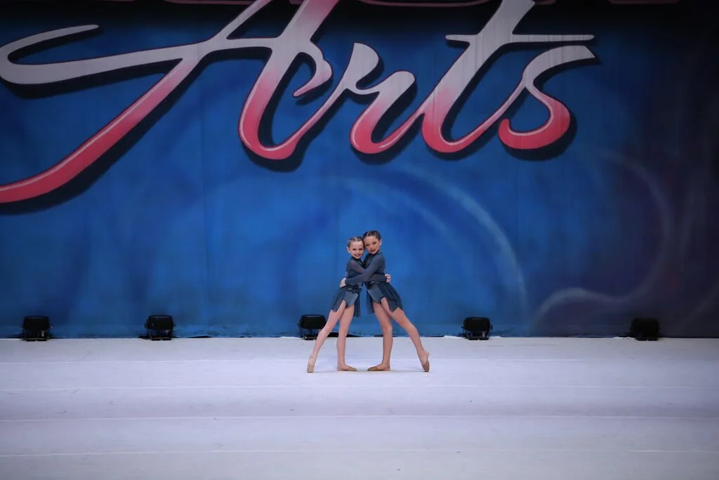 Two young female dancers in costumes performing a dance routine on stage with a large backdrop that reads "Arts."