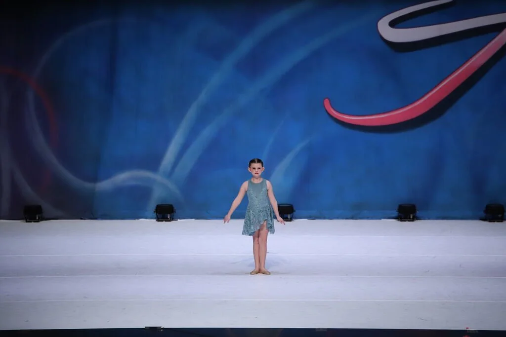 A young girl performing a dance routine on a stage with a blue backdrop and stage lights.