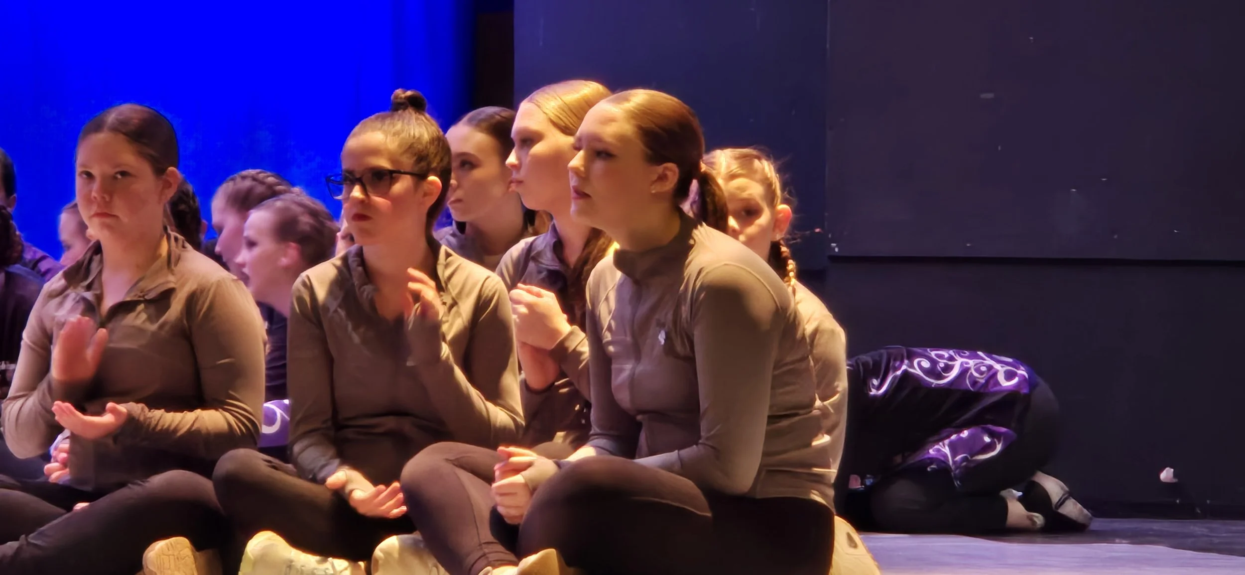 Group of young girls sitting on the floor, watching a performance or event with attentive and serious expressions, in a dimly lit room with purple and blue lighting.