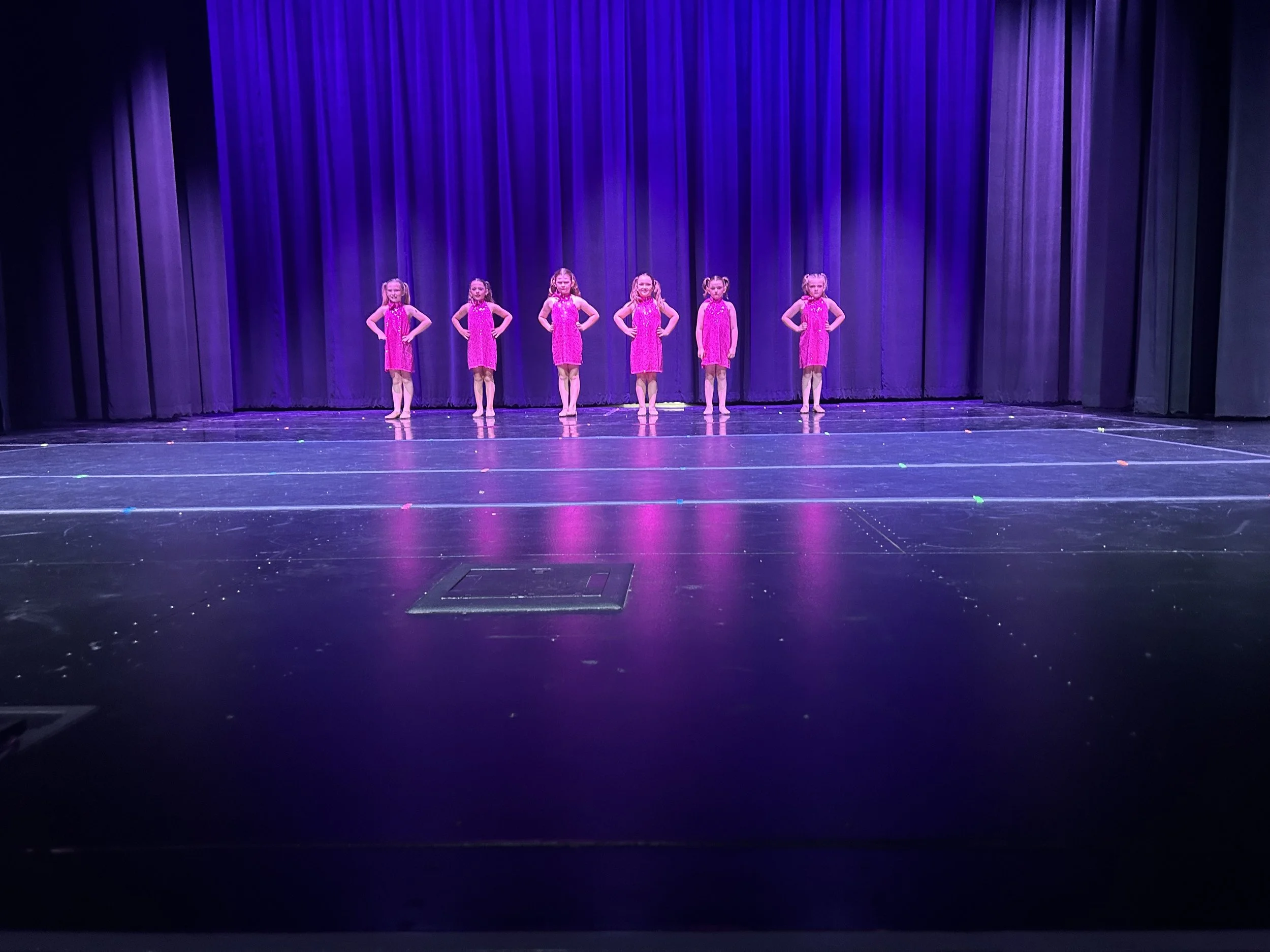 Six young girls in pink costumes standing with hands on hips on a stage with purple curtains.