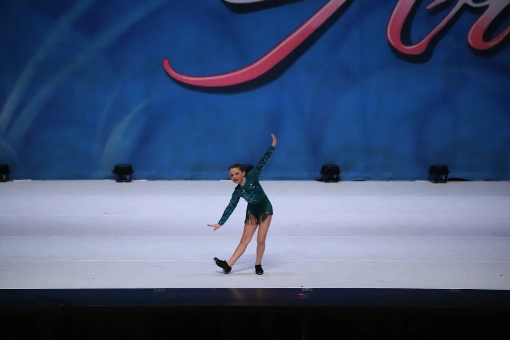 A young female figure skater performing on an indoor ice rink, dressed in a teal sequined costume with black ice skates, with a large blue backdrop featuring part of a red-colored word.
