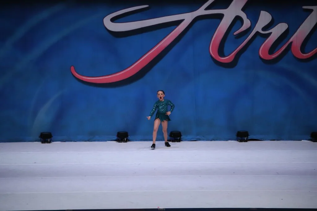 A young girl performing a dance on stage with a blue backdrop and large pink and white lettering.