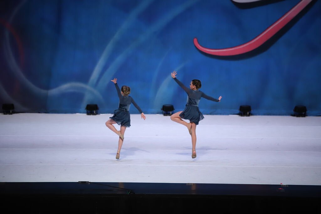 Two young girls in blue dresses performing a ballet dance on stage, standing on one foot with their arms raised, in front of a colorful abstract backdrop.