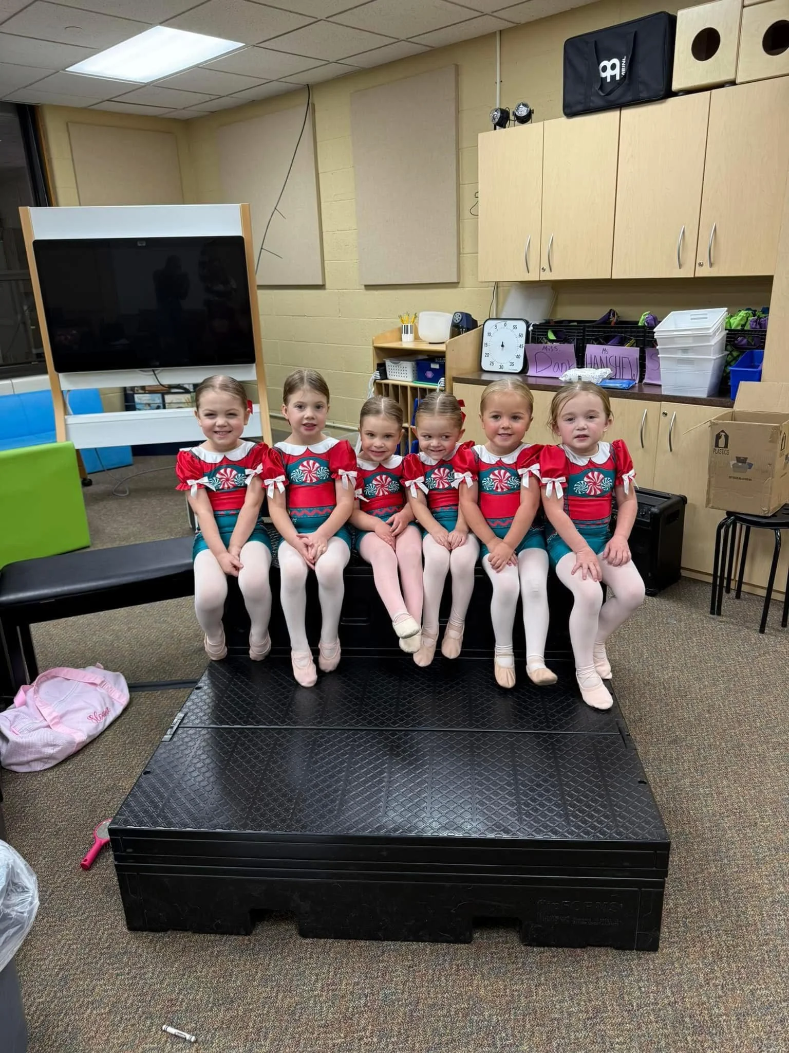 Six young girls sitting on a black platform, dressed in holiday-themed costumes with red and white tops, light-colored tights, and ballet slippers, in a classroom setting with storage cabinets, a large digital screen, and colorful bins in the backgro