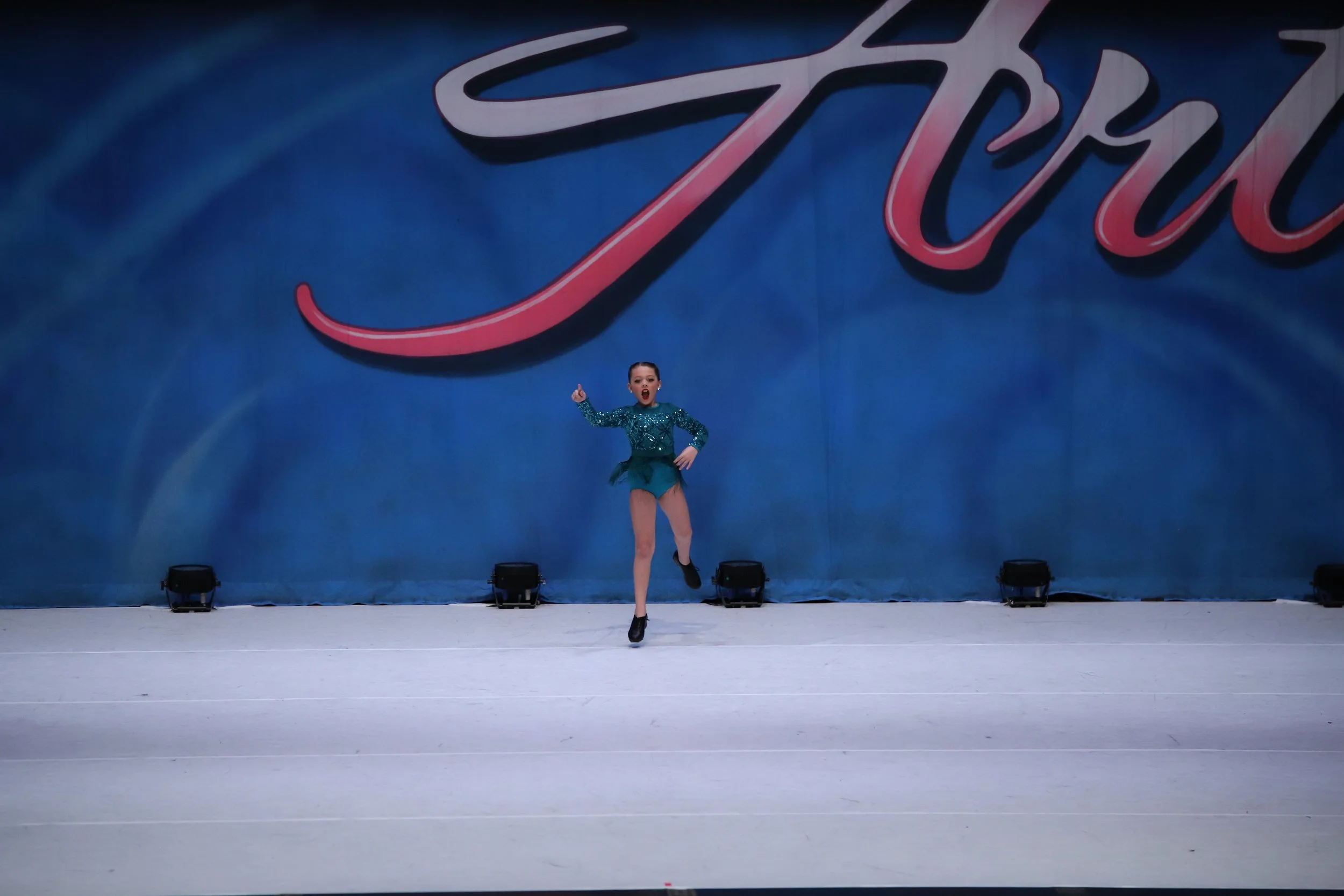 A young female figure skater performing on an ice rink in front of a large blue backdrop with the word 'Star' in stylized red and white lettering. She is mid-jump, wearing a teal costume with sequins and black ice skates.