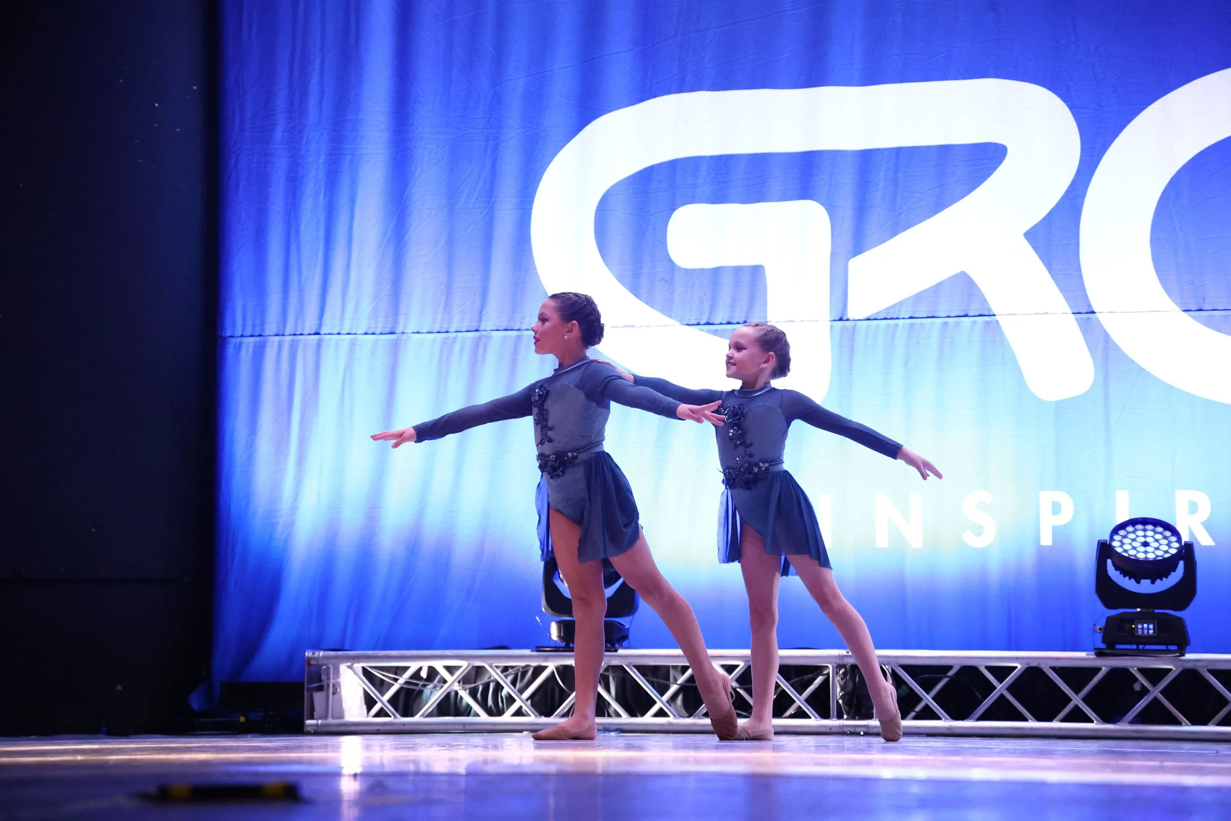 Two young female dancers in matching dark blue costumes perform on stage during a dance competition, with a large illuminated backdrop behind them.