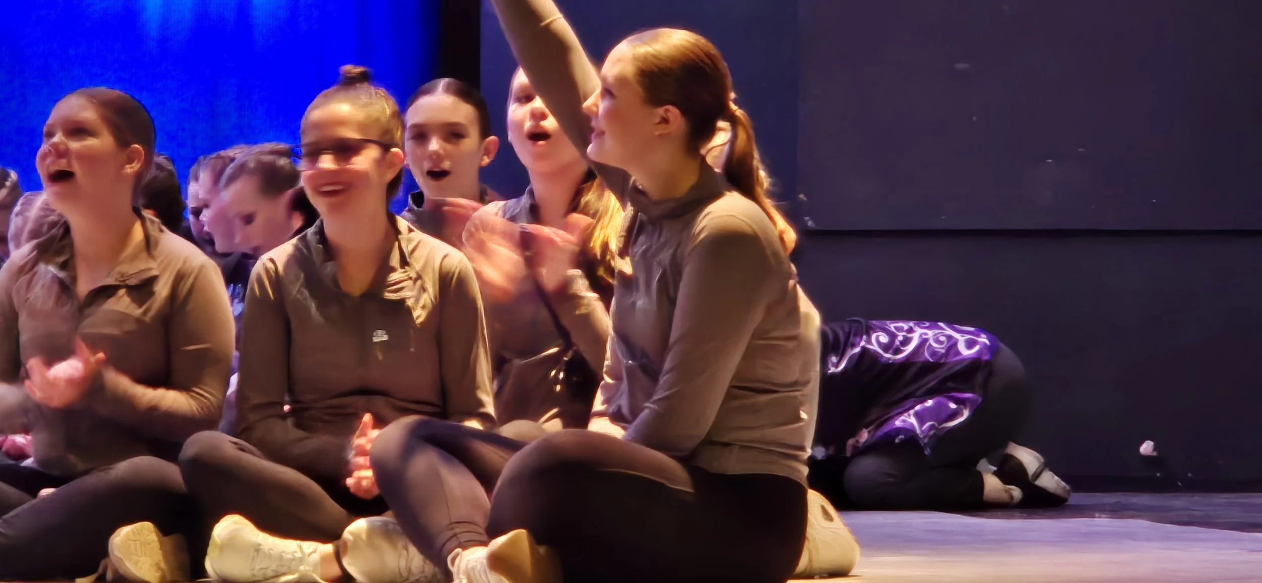 Group of young girls sitting on the stage floor, laughing and clapping, during a performance or event, with some of them showing excitement and joy.