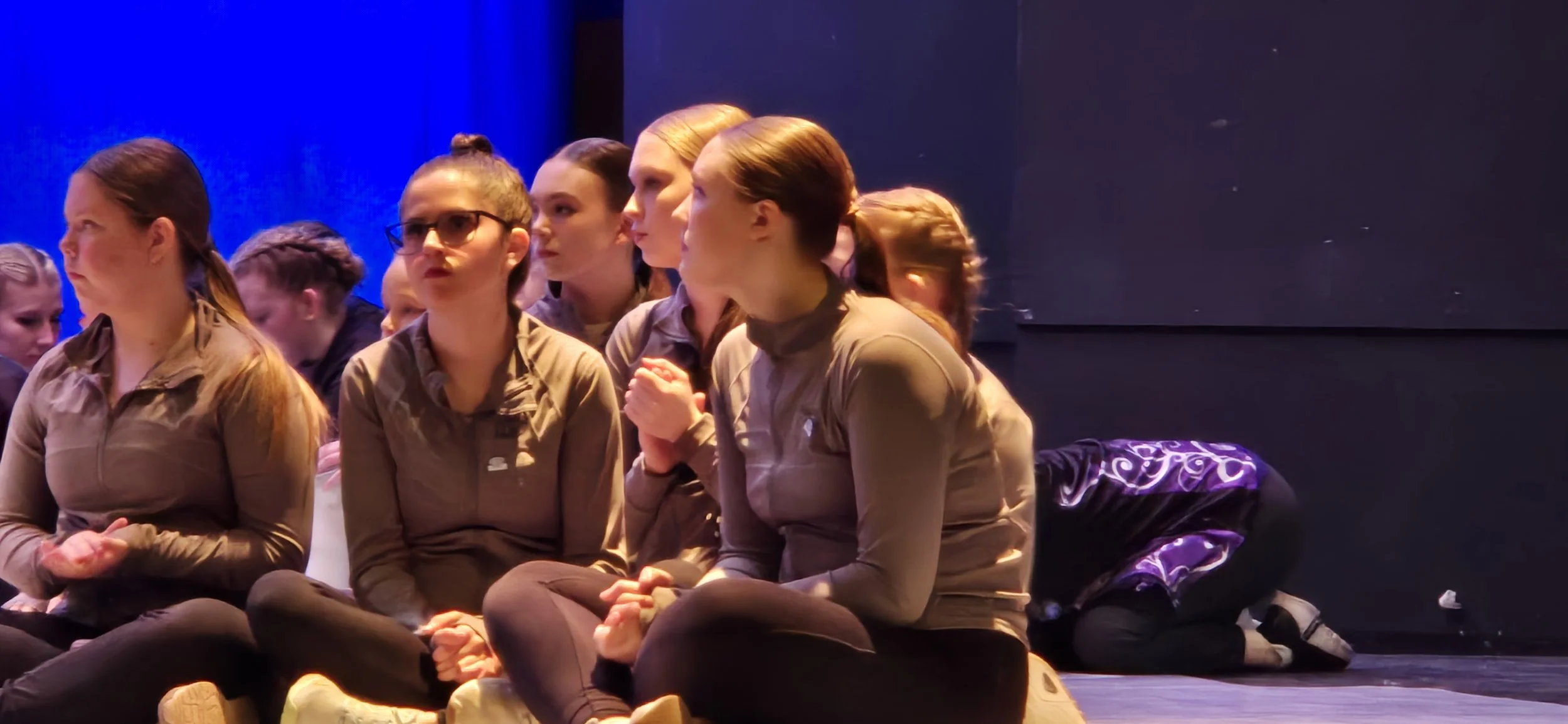 A group of women sitting on the floor, with one woman kneeling in the background, under stage lighting.