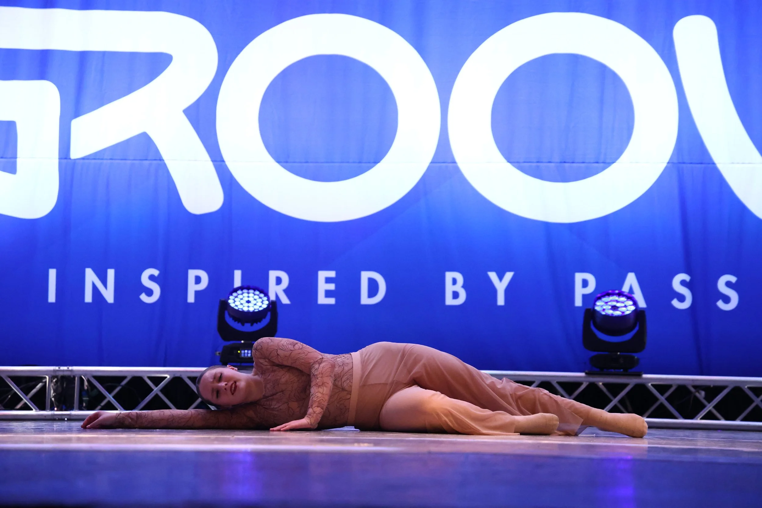 A female dancer lying on her side on a stage, performing a dance move with her left arm extended and her head resting on her arm, against a backdrop with a large logo and the words 'INSPIRED BY PASS' illuminated by stage lights.