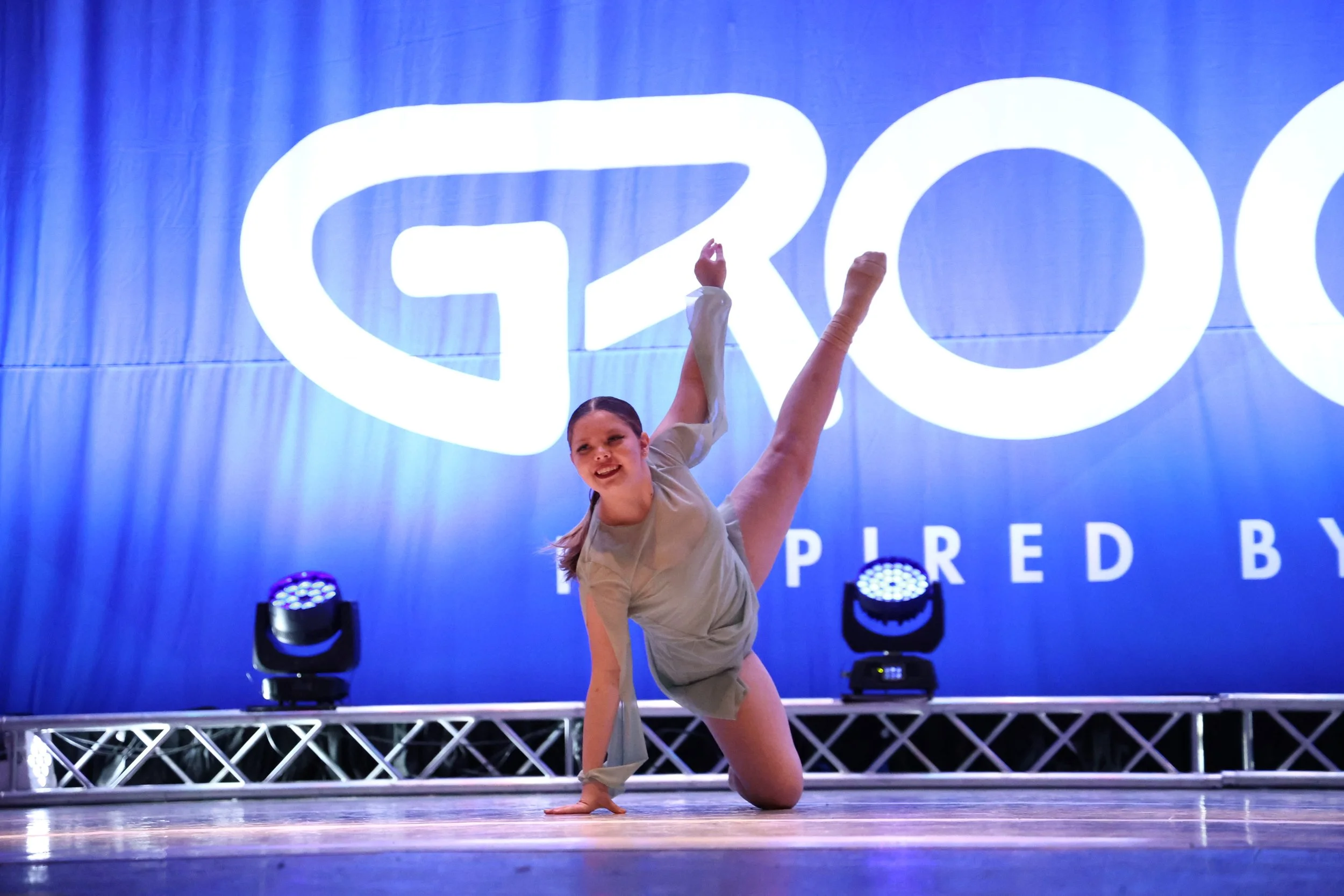 Young female dancer performing a pose on stage during a dance competition, with a blue backdrop that reads 'GRO' and 'SPRED BY' in the background.