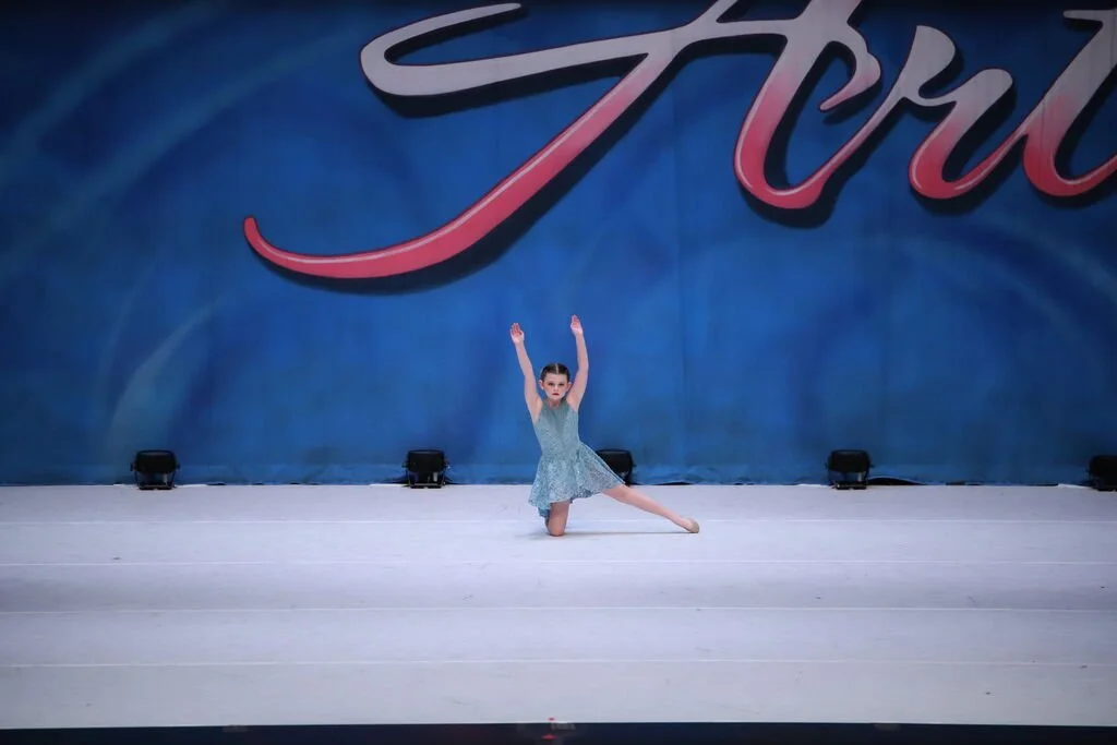 A young girl performing a dance on stage in front of a large blue backdrop with the word 'Tro' visible. She is wearing a light blue dress and is striking a pose with her arms raised and one leg extended to the side.
