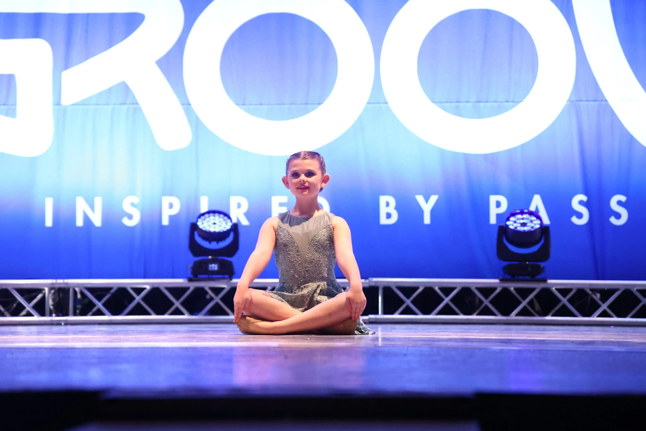 Young girl sitting cross-legged on stage during a dance or performance, with a large blue backdrop displaying the word 'GROOVE' and the phrase 'INSPIRED BY PASS' behind her.