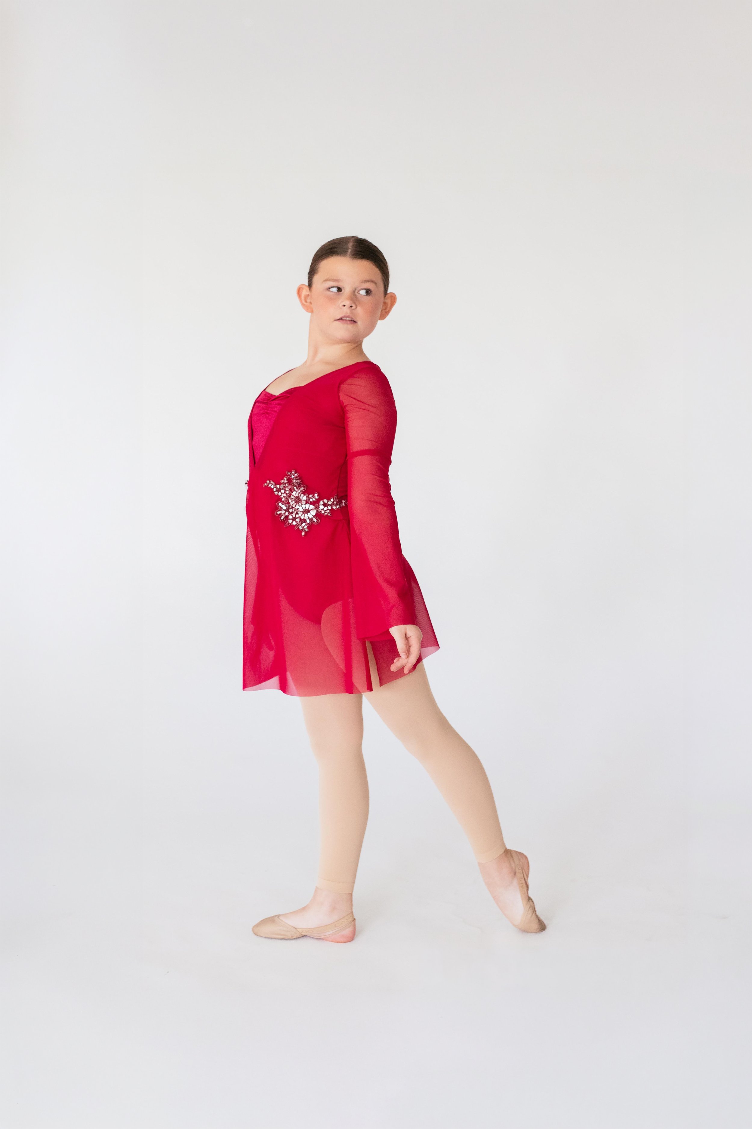 A young girl practicing ballet dance in a red dress and beige ballet shoes against a plain white background.