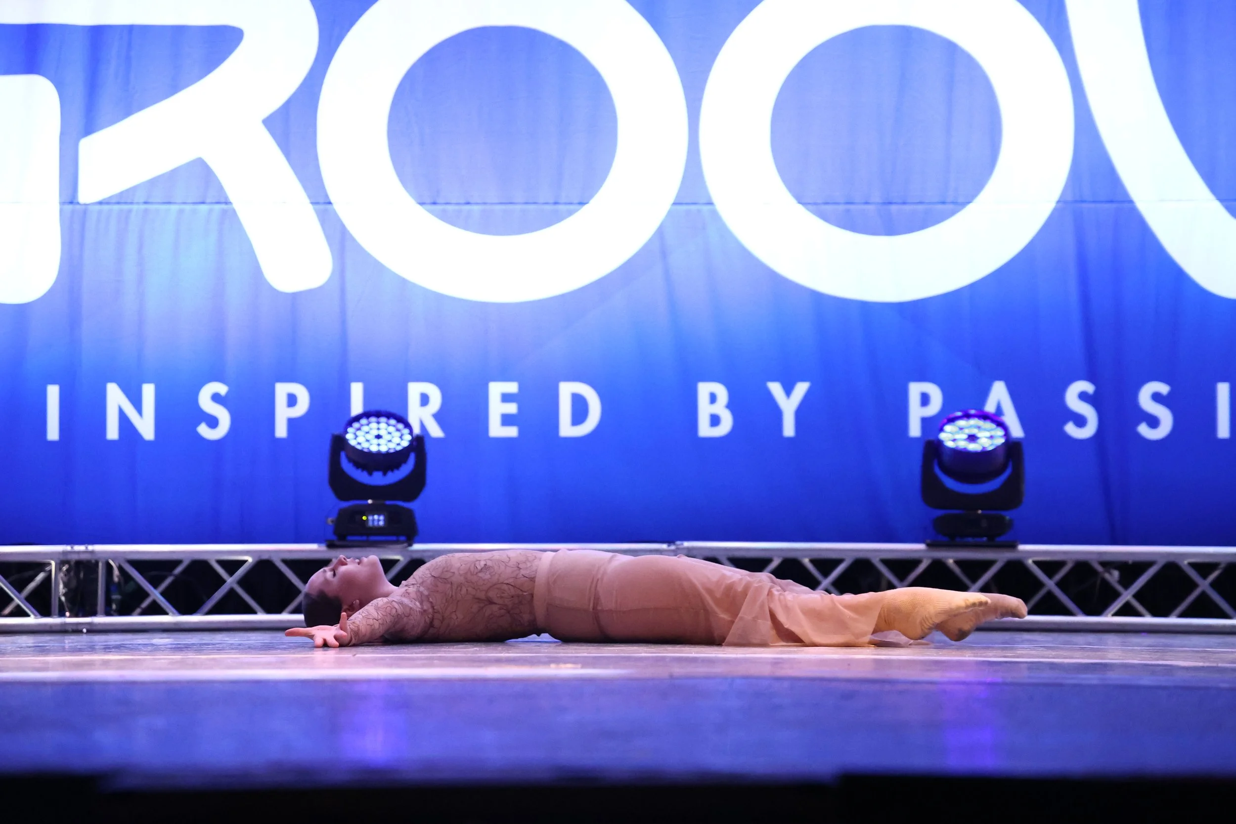 A young girl performs a dance move on stage lying flat on her back with arms extended outwards, in front of a large blue backdrop with white text and stage lights.