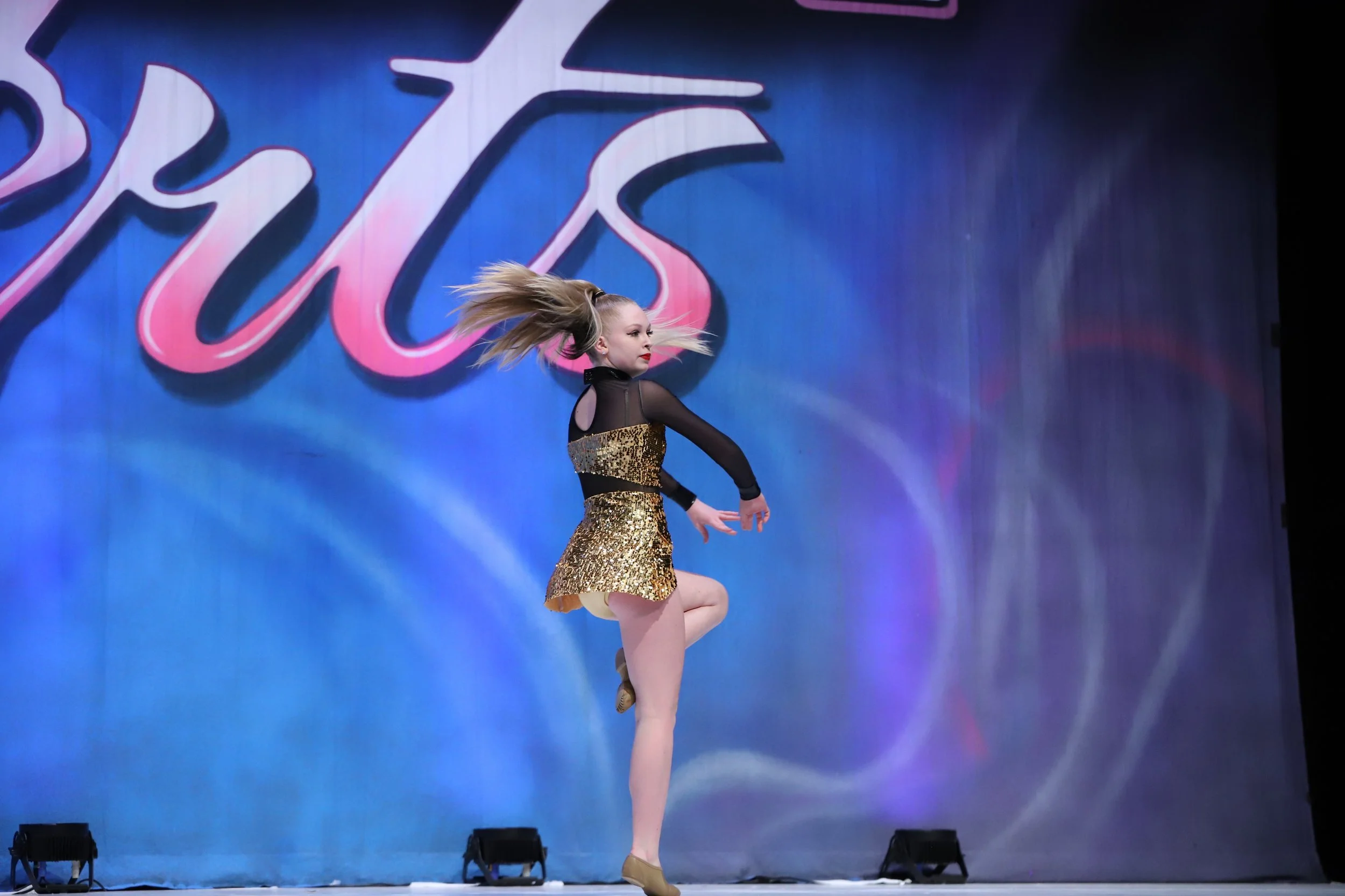 Young female dancer mid-performance on stage, wearing a black and gold dance costume, with her hair flying and one leg raised.