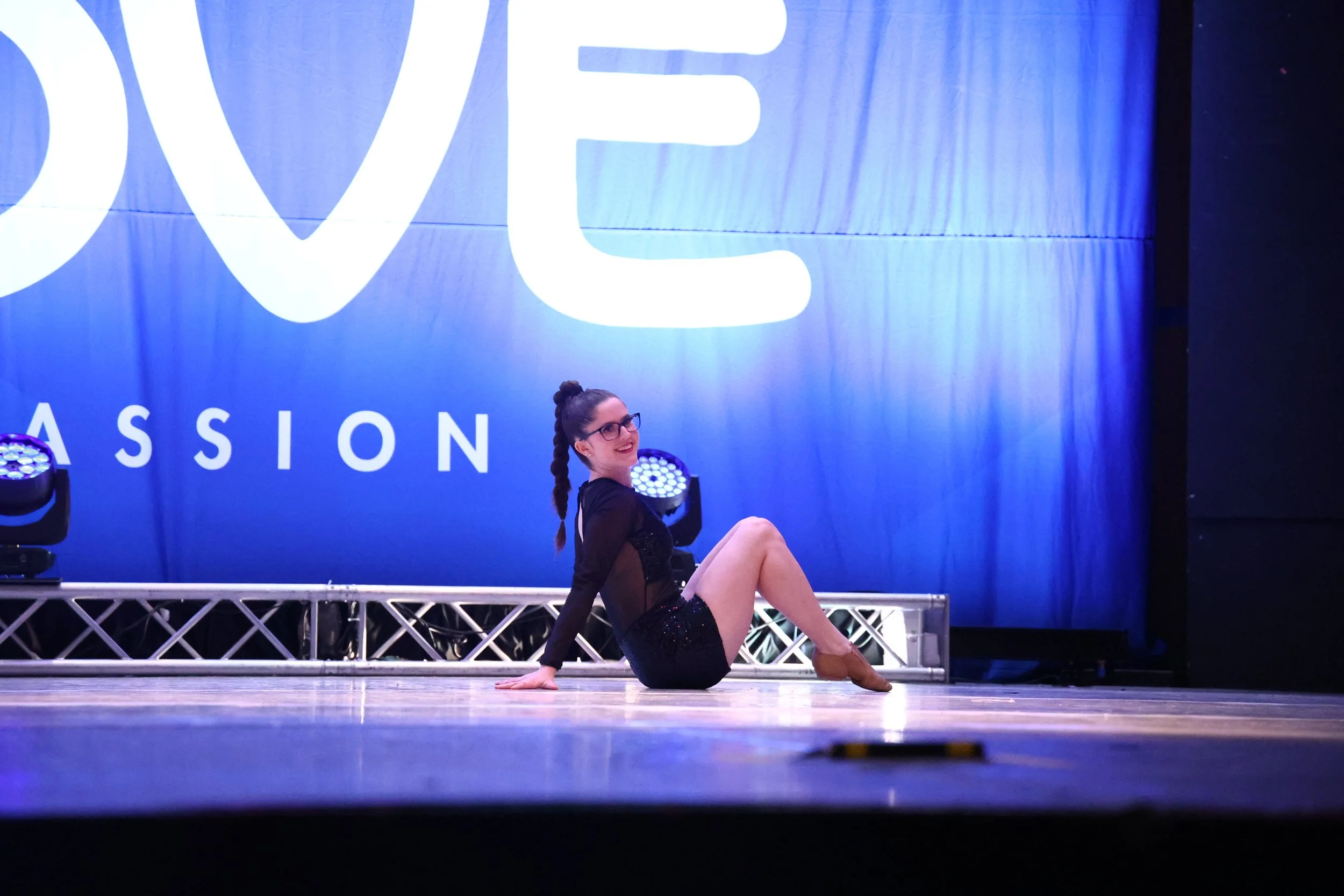 A woman in a black dance costume and glasses sitting on a stage with a blue background that says 'LOVE' and 'PASSION', smiling at the camera.