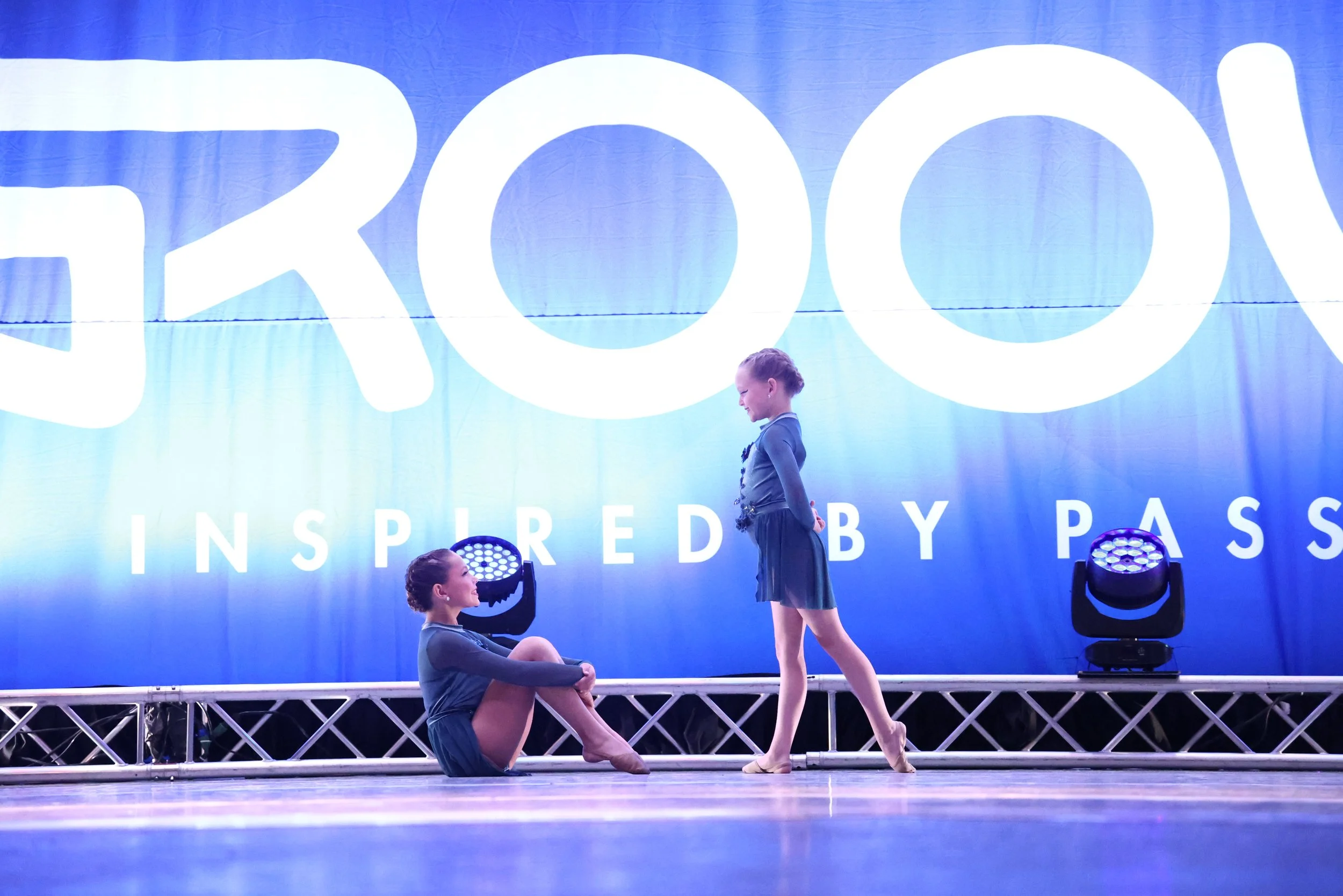Two young girls in dance costumes performing on stage with a large blue backdrop that reads 'FROG INSPIRATION PAS'. One girl is sitting on the floor with legs crossed, looking at the other girl who is standing in a pose, both wearing matching dance o