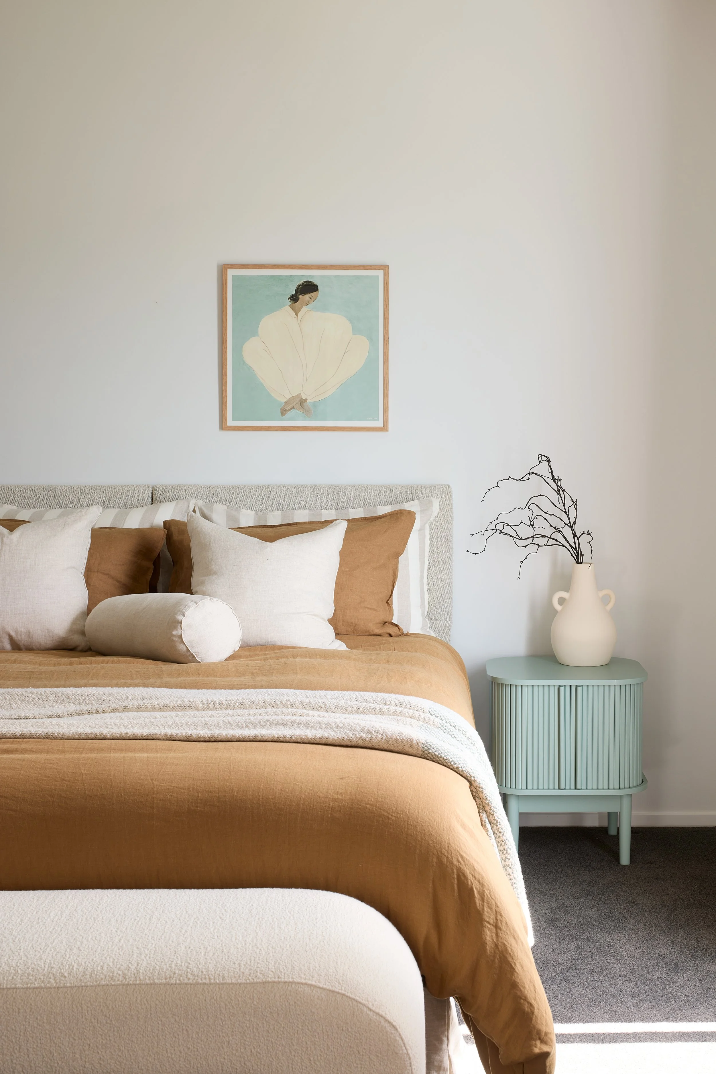 A cozy bedroom with a beige bedspread, multiple pillows, and a framed artwork of a woman on the wall. Next to the bed is a mint green nightstand with a vase and branches, on light gray carpeted flooring.