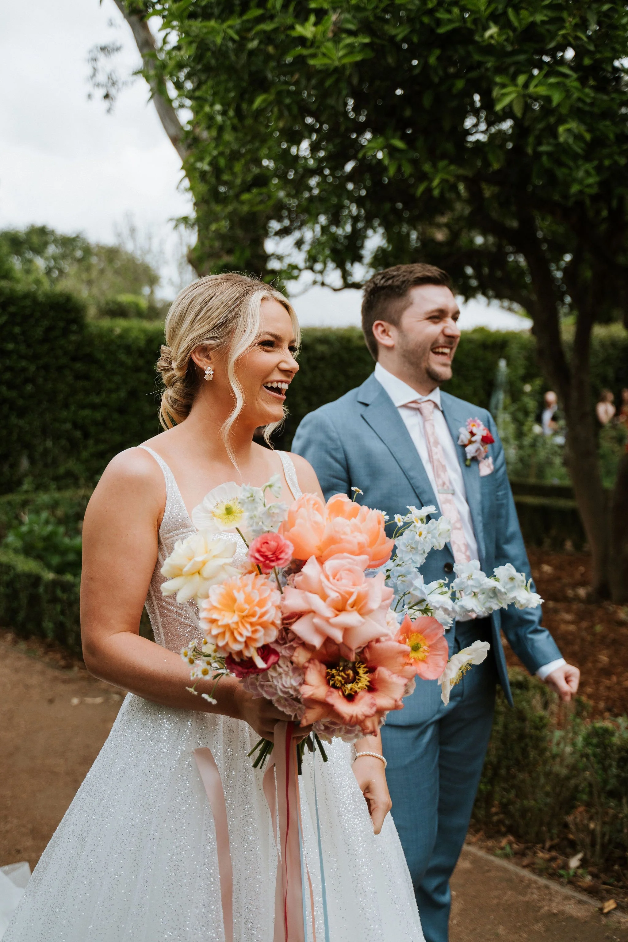 A bride and groom smiling during their outdoor wedding ceremony. The bride holds a colorful bouquet of flowers, and the groom wears a blue suit with a floral boutonniere. They are walking outdoors with trees and greenery in the background.
