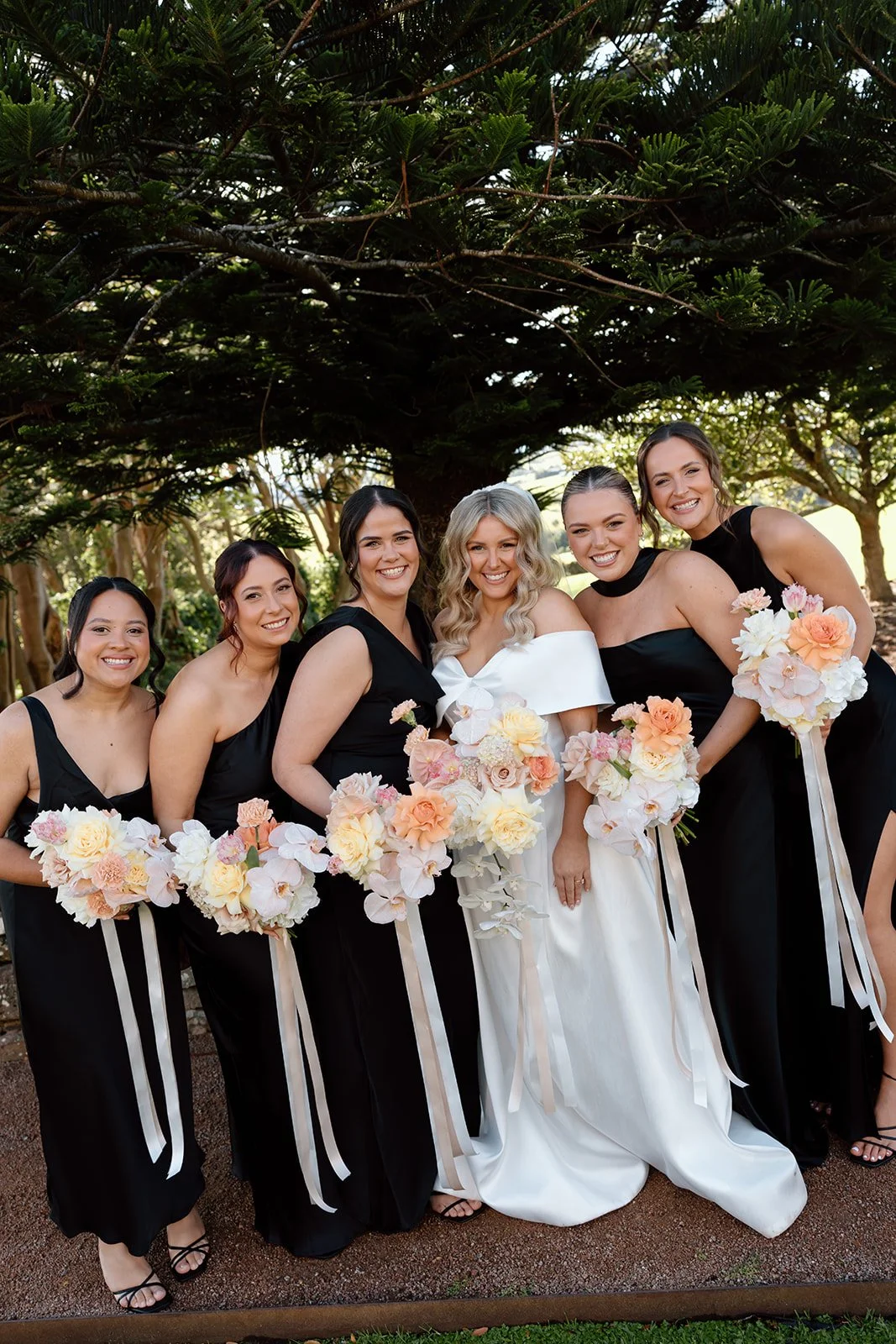 Bride in a white gown and five bridesmaids in black dresses, holding bouquets with peach, pink, and white flowers, standing outdoors under a large tree.