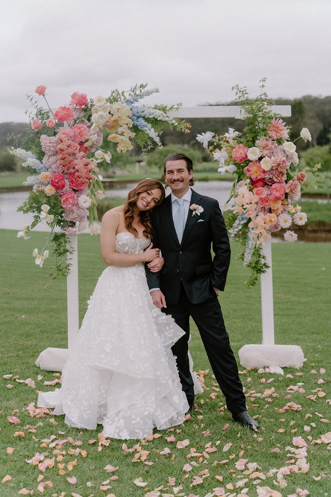 A bride and groom standing under a large floral wedding arch outdoors on a grassy area with rose petals scattered on the ground, smiling and posing for a photo.