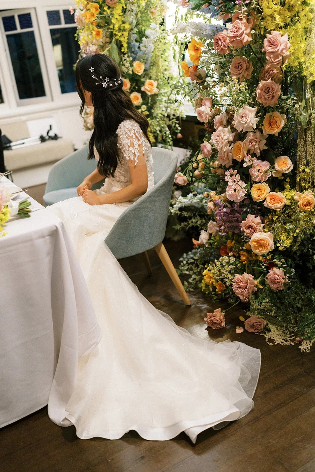 A bride in a white wedding gown with lace details sitting at a table decorated with flowers and a large floral arrangement in the background.