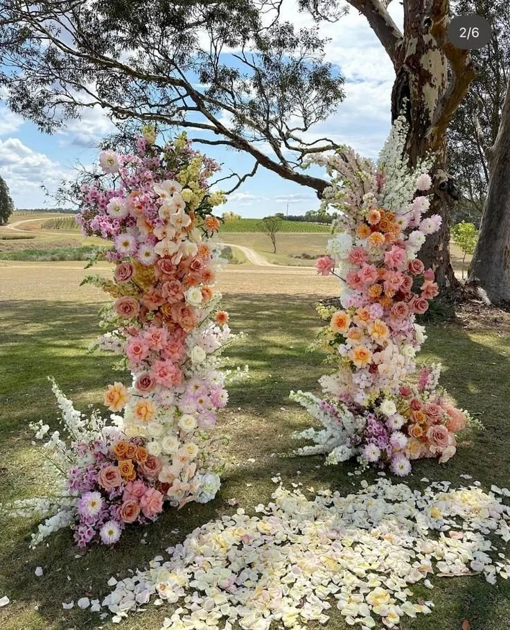 Outdoor wedding arch decorated with pink, white, and peach flowers, with flower petals scattered on the ground, under a tree in a rural landscape.