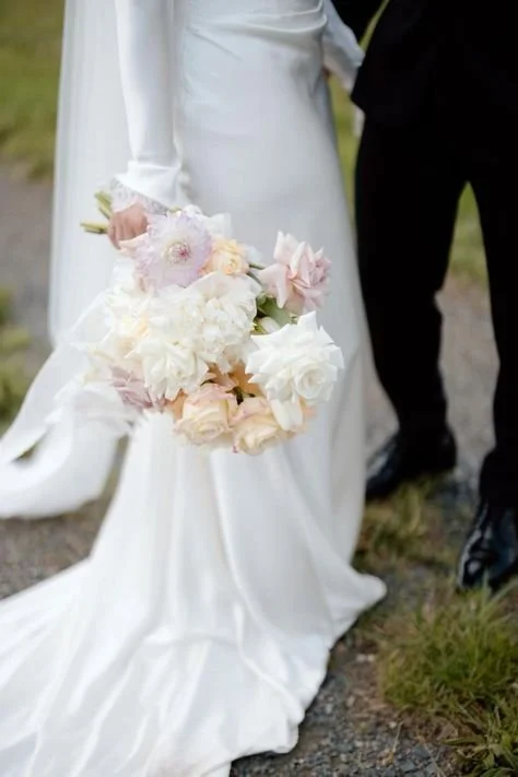 Bride holding a bouquet of white and pale pink flowers in her right hand, wearing a long white wedding dress, with a groom standing next to her in black pants and shoes.